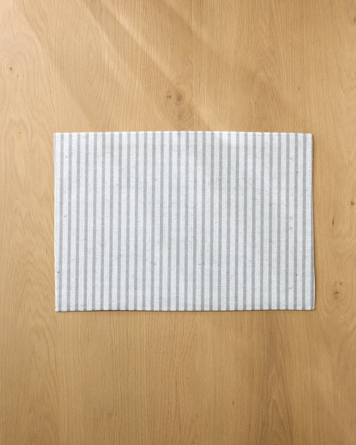 A rectangular Stone Stripe placemat by Kailee Wright, featuring light gray and white stripes, is centered on a light wood table with a natural grain pattern.