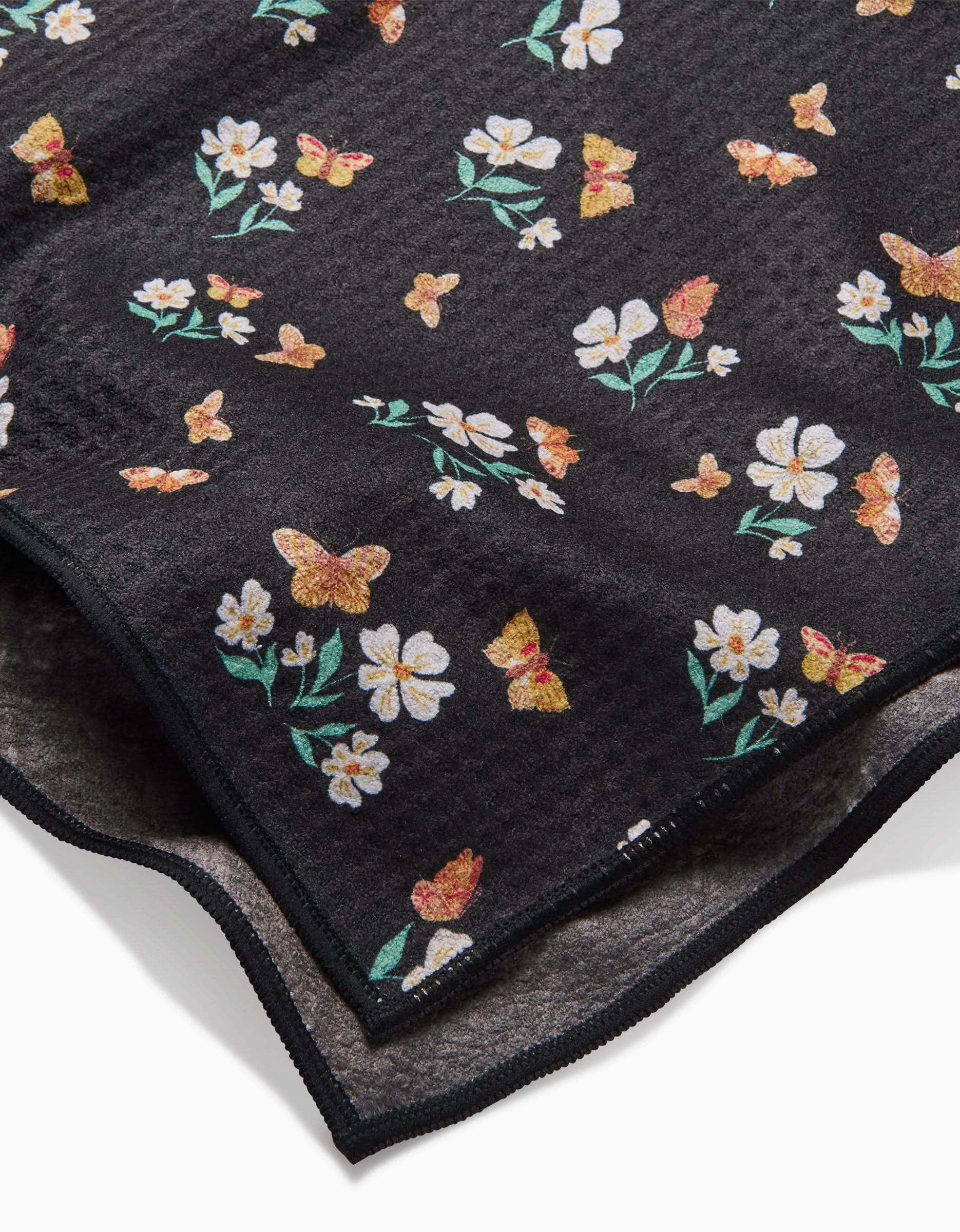 A close-up of Danelys Sidron’s Butterfly Garden towel displays black Geoweave fabric with vibrant floral and butterfly prints, featuring yellow, white, and orange accents. The ultra-absorbent texture and stitched edges are visible.