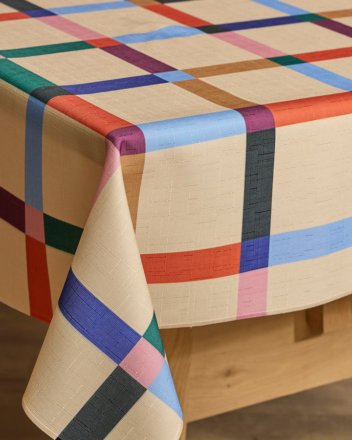Jennifer Bouron's Cool Grid tablecloth, featuring a beige, eco-friendly design with vibrant blue, red, green, pink, purple, and black plaid stripes, is draped over a wooden table.