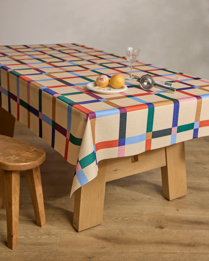 A Cool Grid tablecloth by Jennifer Bouron covers a wooden table set with two onions and a garlic bulb on a plate, a metal ice cream scoop, an empty glass, and stands next to a wooden stool on a light wood floor.