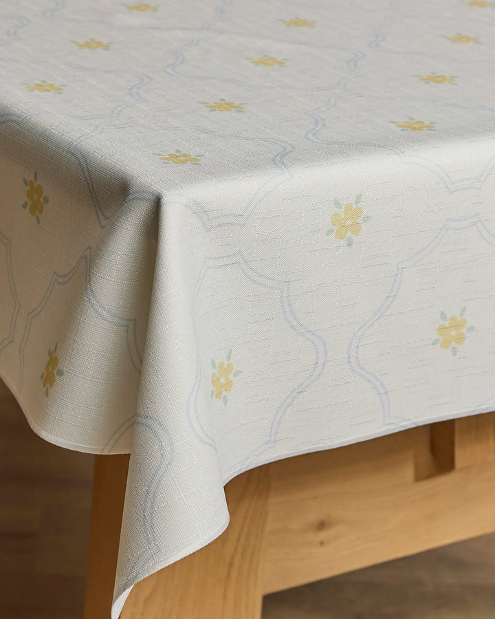 A close-up of a wooden table draped with Jasmine Blooms Designs’ Dainty Blooms tablecloth, featuring a lightweight fabric with a subtle geometric pattern and small yellow floral motifs.