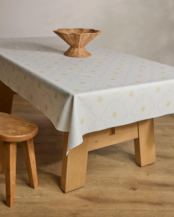 A wooden table draped with the "Dainty Blooms" lightweight, spill-resistant yellow floral tablecloth from Jasmine Blooms Designs holds an empty wicker basket. A wooden stool sits next to it on a wooden floor.