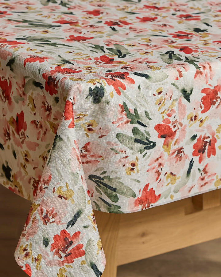 A rectangular wooden table is draped with the Painted Garden tablecloth by Melissa Johnson, featuring a sustainable white fabric adorned with red, green, yellow, and pink floral prints.