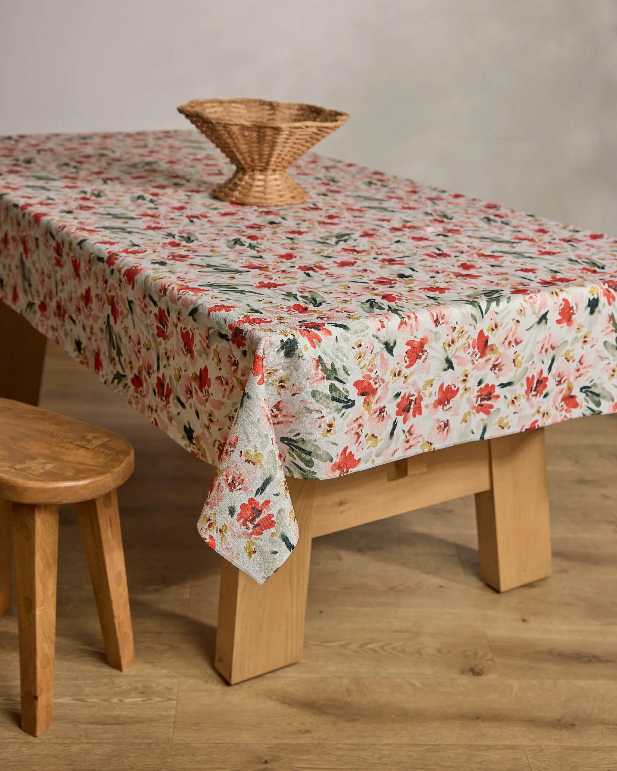 A wooden table is covered with Melissa Johnson's Painted Garden sustainable, floral-patterned tablecloth in red, green, and white, topped with a woven basket centerpiece; a wooden stool stands beside it on the light wood floor.