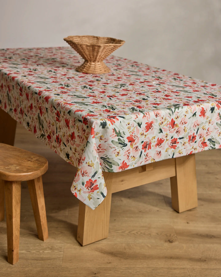 A wooden table is covered with Melissa Johnson's Painted Garden sustainable, floral-patterned tablecloth in red, green, and white, topped with a woven basket centerpiece; a wooden stool stands beside it on the light wood floor.
