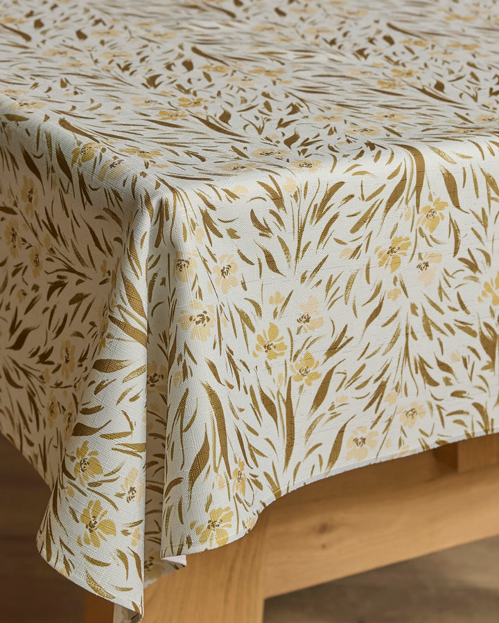 The Melissa Johnson Yellow Floral tablecloth, cream-colored with a gold and brown floral and leaf design, is spill-resistant and drapes elegantly over your wooden table.
