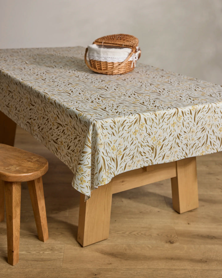 A wooden table is covered with a Melissa Johnson "Yellow Floral" spill-resistant tablecloth. On top sits a small woven basket with folded white napkins, and a wooden stool is placed beside it on a light wood floor.
