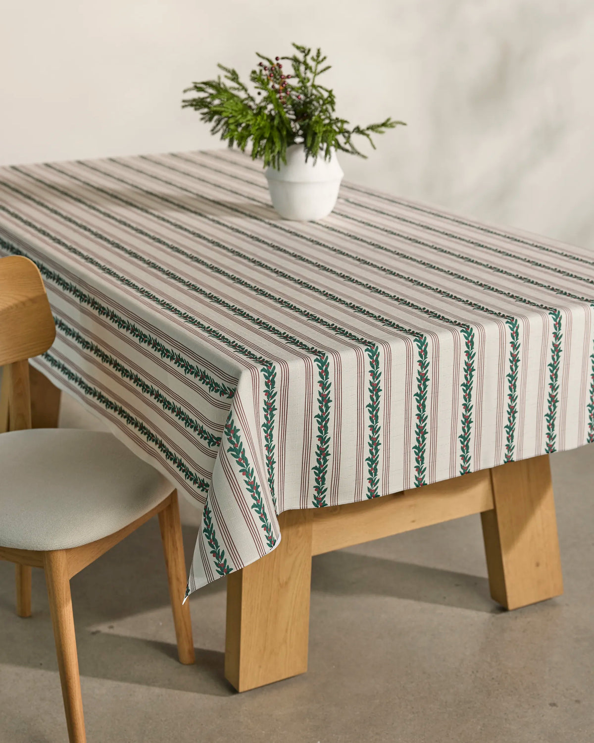 A wooden table adorned with Geometry’s Berry Grove lightweight, striped tablecloth featuring green and red leaf patterns, paired with a wooden chair with a white cushion, and accented by a small plant in a white pot as the centerpiece.