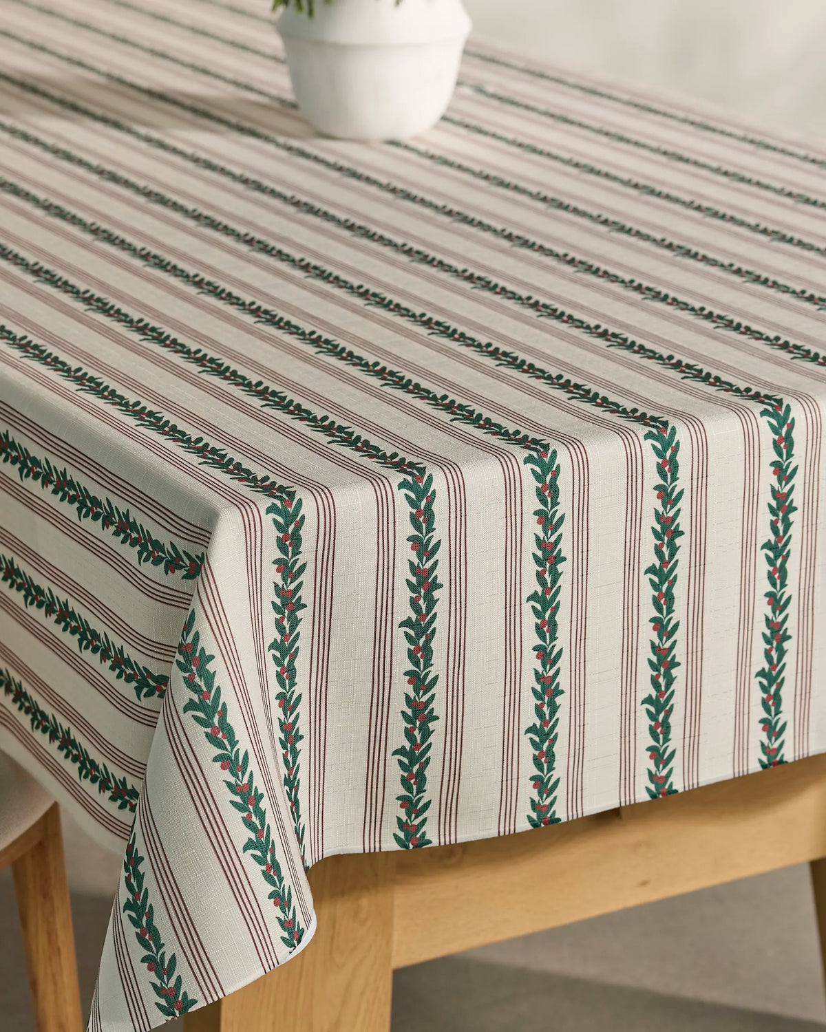 A close-up of a table covered with Geometry’s Berry Grove tablecloth—a lightweight, spill-resistant beige cloth featuring green and brown vertical stripes adorned with leaf patterns—topped with a white potted plant.