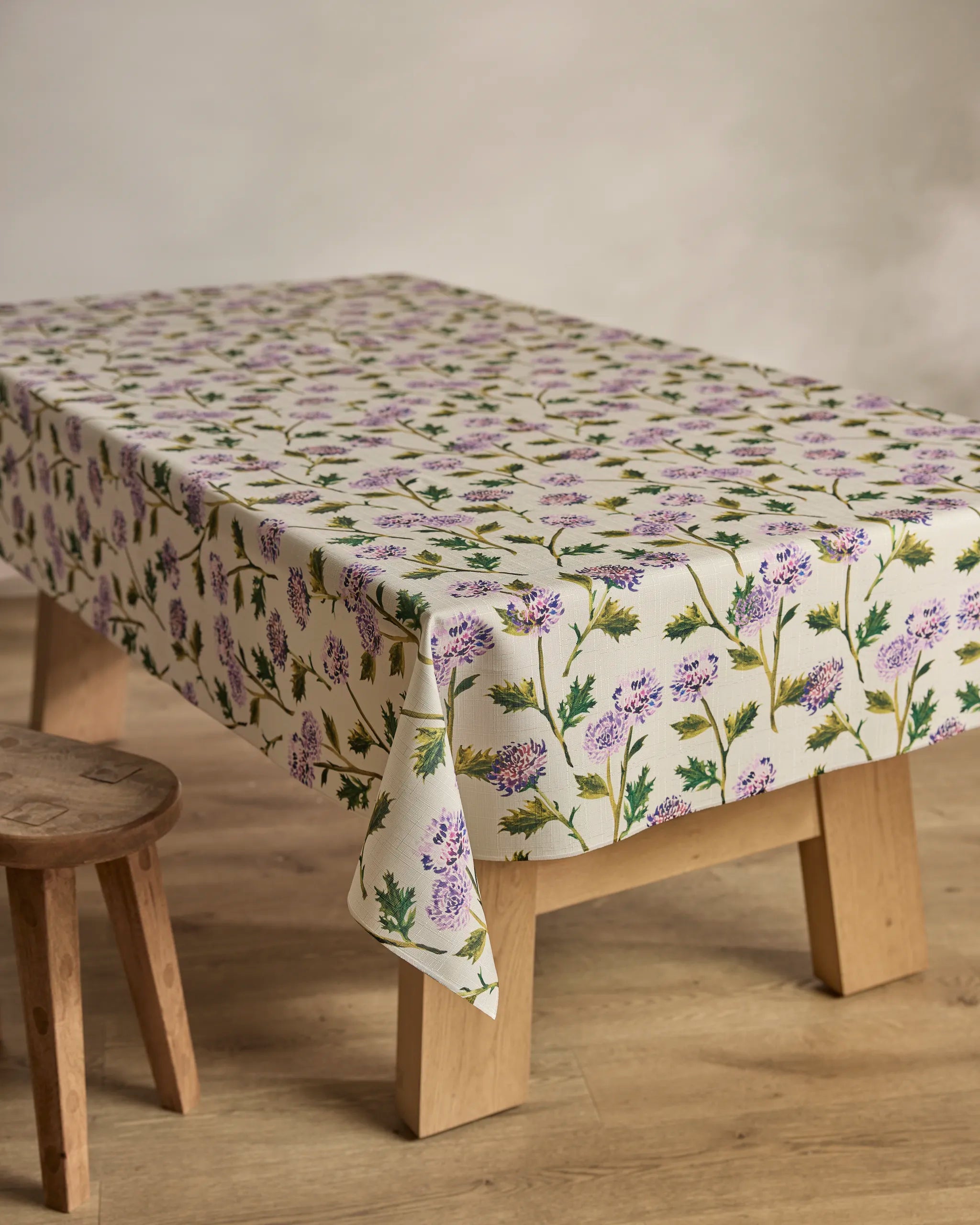 The Geometry "Blooming Thistle" eco-friendly cream tablecloth with a purple and green floral pattern covers a wooden table beside a wooden stool, all set on a light wood floor with a neutral backdrop.