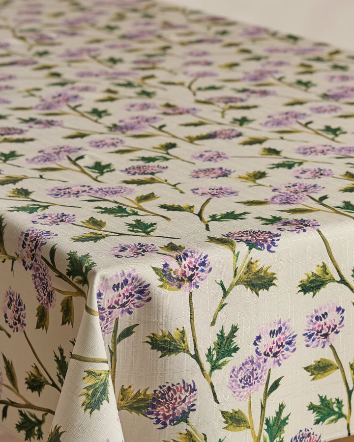 The Geometry Blooming Thistle rectangular tablecloth, in a cream shade with purple thistle flowers and green leaves, is spill-resistant and drapes elegantly over your table.