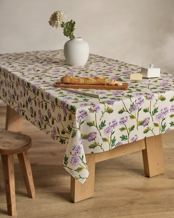 A wooden table is adorned with Geometry's Blooming Thistle eco-friendly purple floral tablecloth, topped with a cutting board holding a baguette, butter dish, and a white vase with a single white flower. A wooden stool sits nearby on the floor.