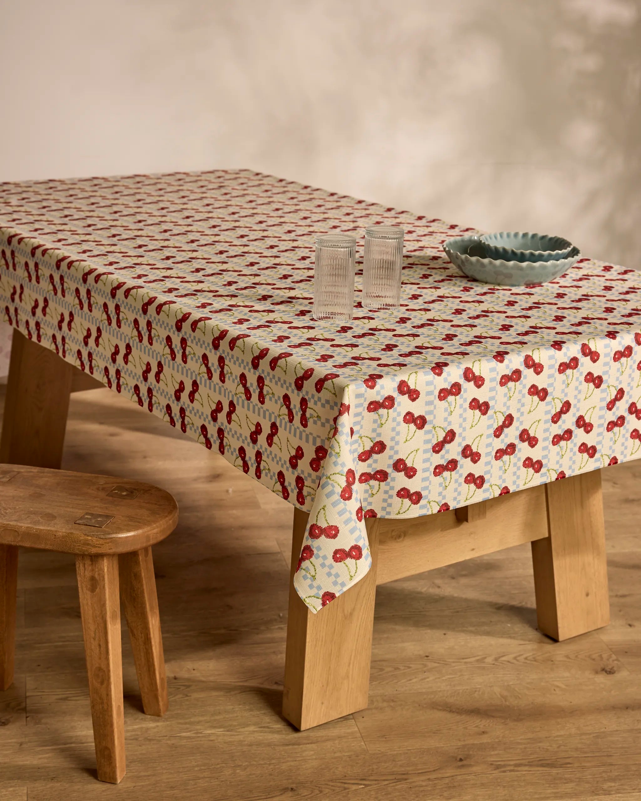 A wooden table is topped with Geometry's Cherry Cola spill-resistant tablecloth, two ribbed glass tumblers, and a scalloped blue bowl. A wooden stool stands nearby on a light wood floor with soft wall shadows.