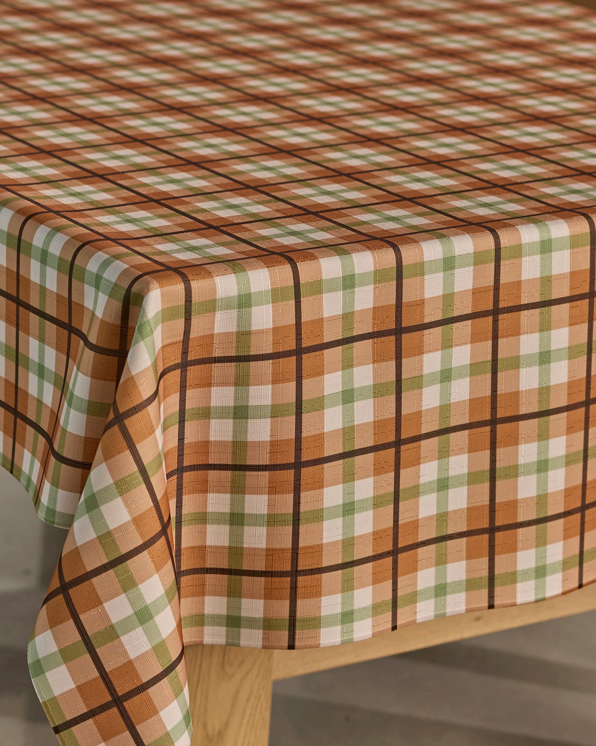 A close-up of a wooden table topped with Geometry’s Country Kitchen tablecloth, featuring lightweight brown, green, and white plaid checks.