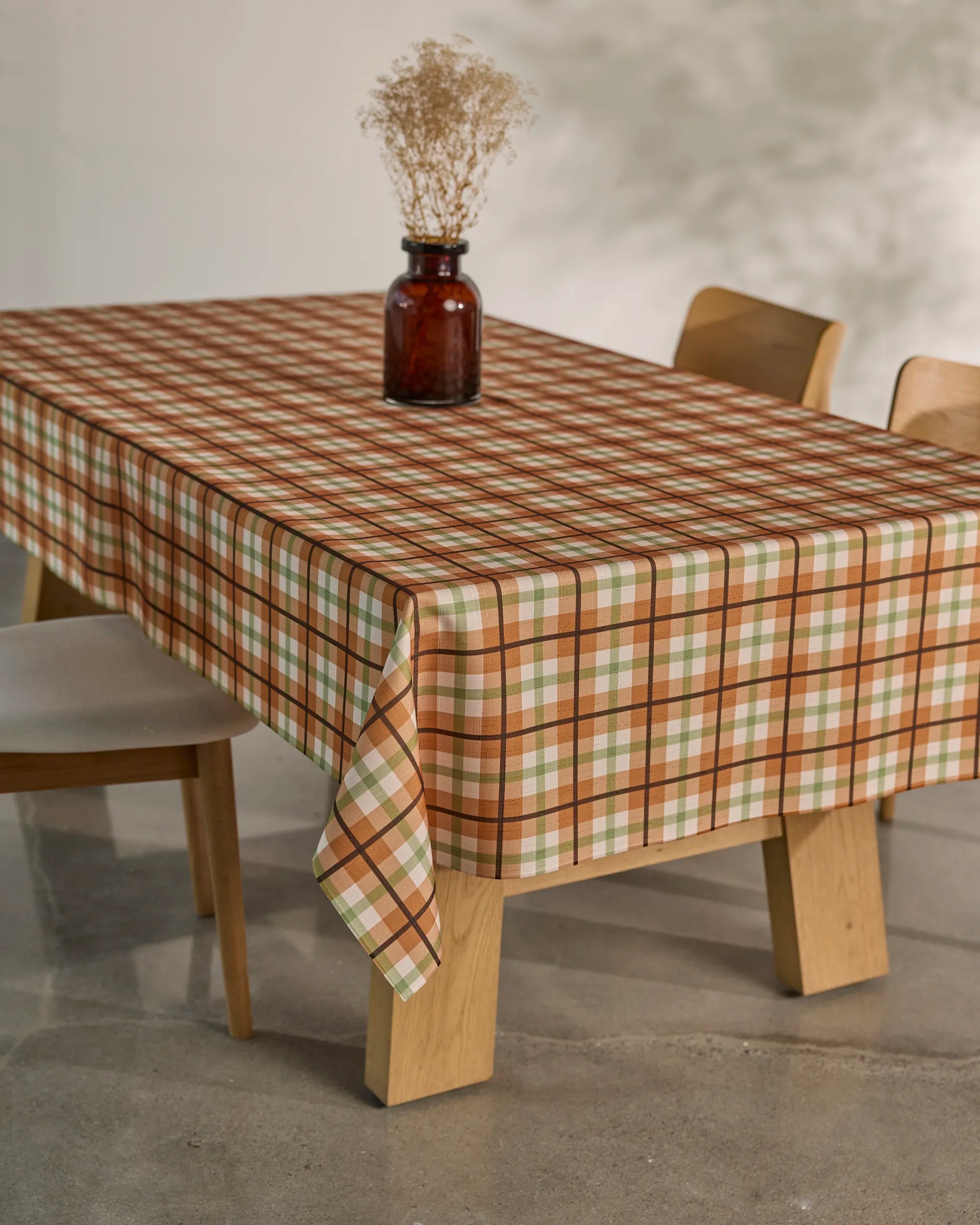 The Country Kitchen dining table by Geometry features a plaid spill-resistant tablecloth in brown, green, and cream, topped with a brown glass vase of dried flowers. Two wooden chairs with white seats accompany the setup.