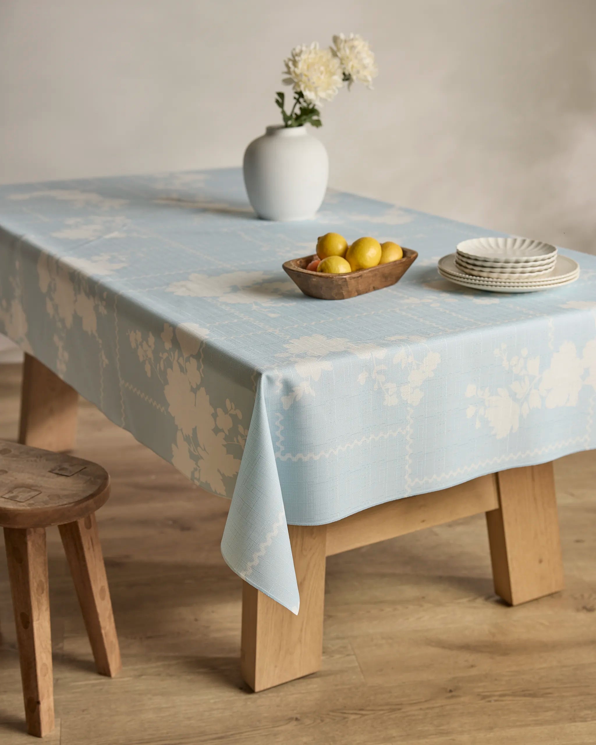 A wooden table draped with Geometry's "Floral Whispers" tablecloth, holding a white vase of flowers, a wooden bowl of lemons, and stacked white plates; a wooden stool sits nearby.