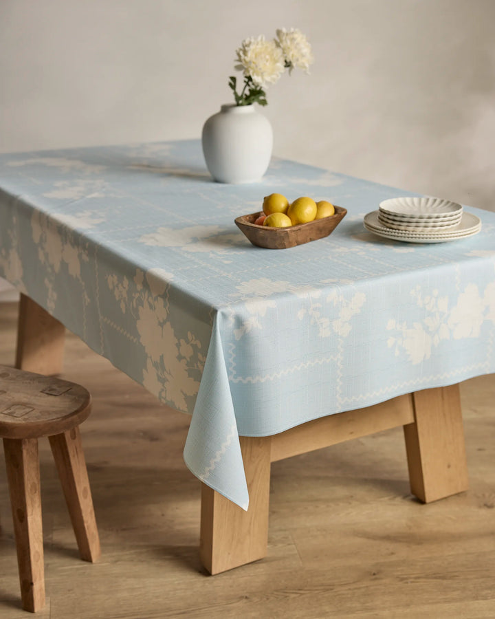 A wooden table draped with Geometry's "Floral Whispers" tablecloth, holding a white vase of flowers, a wooden bowl of lemons, and stacked white plates; a wooden stool sits nearby.
