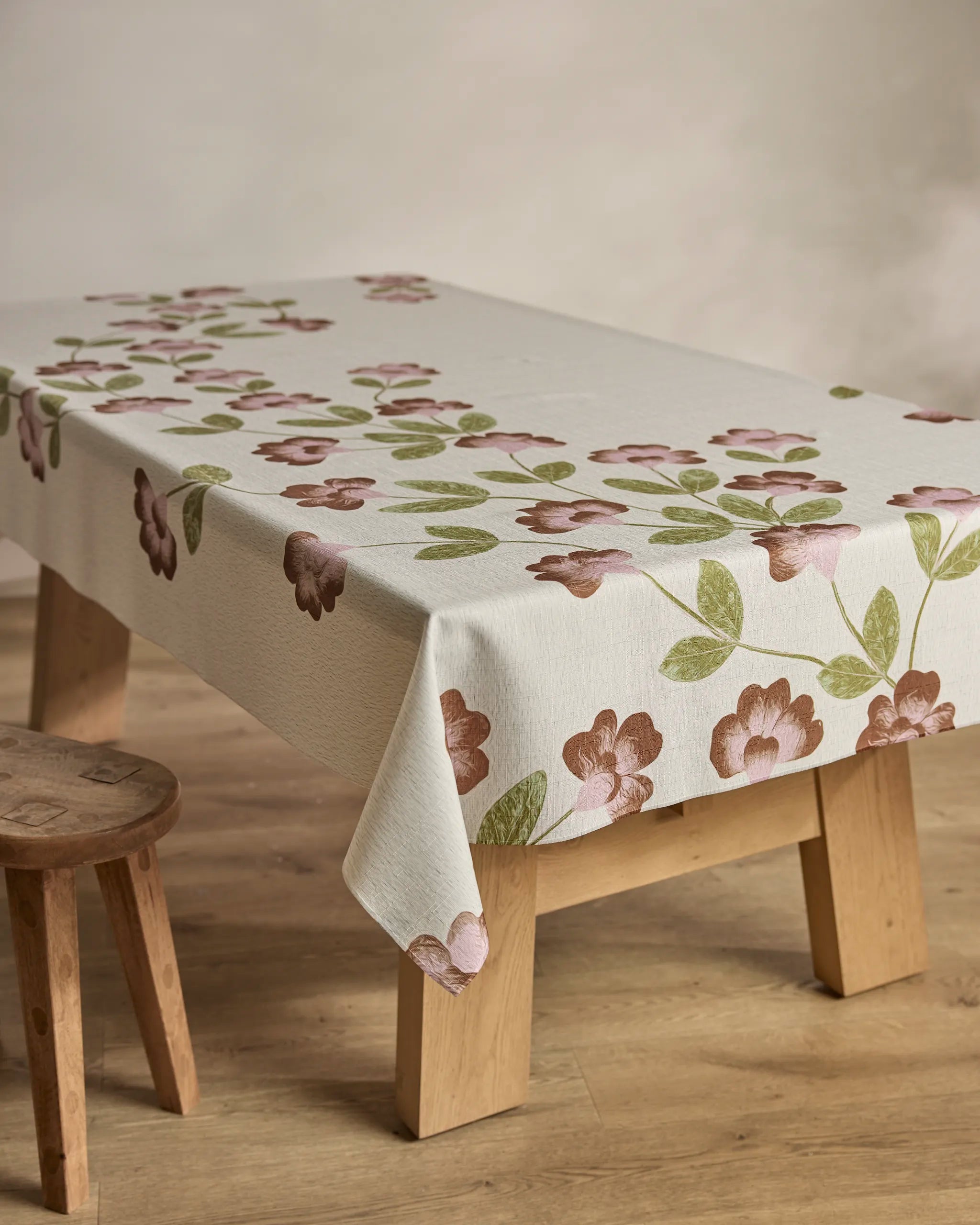 A wooden table is topped with Geometry’s "Flower Tower" spill-resistant tablecloth, featuring pink and brown flowers with green leaves. A wooden stool sits beside the table on a light wood floor, creating a cozy, practical setting.