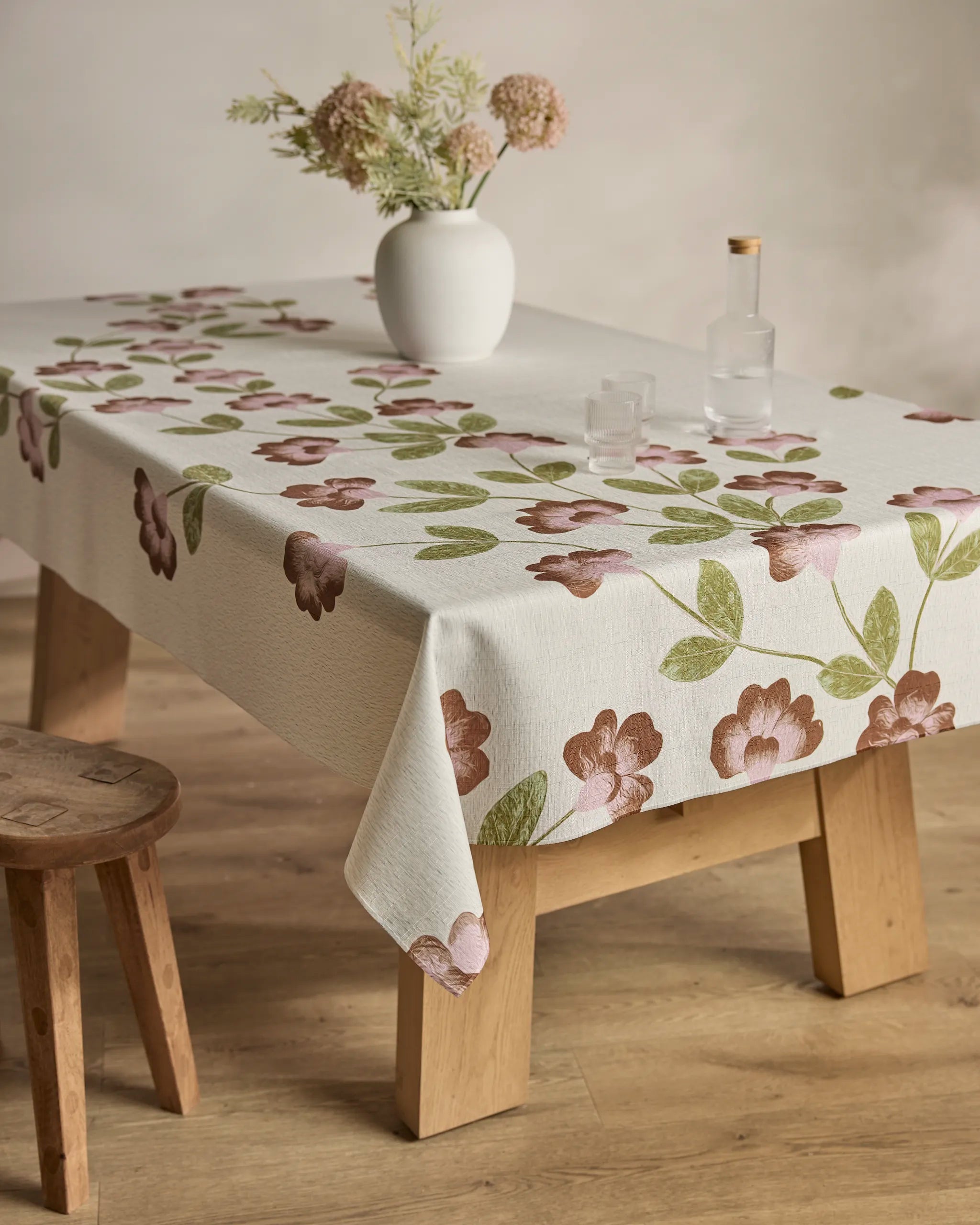 The Flower Tower tablecloth by Geometry, featuring a pink and green floral pattern, covers a wooden table set with a white vase of dried flowers, two clear glasses, and a clear bottle. A wooden stool stands next to the table.