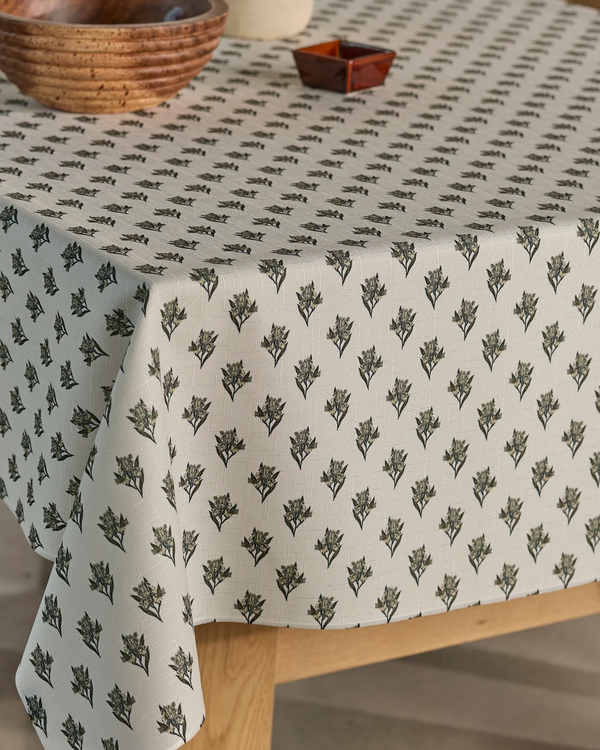 The round table is draped with Geometry’s Foraged, a lightweight cream tablecloth adorned with small green floral motifs, and topped with wooden bowls and a square dish.