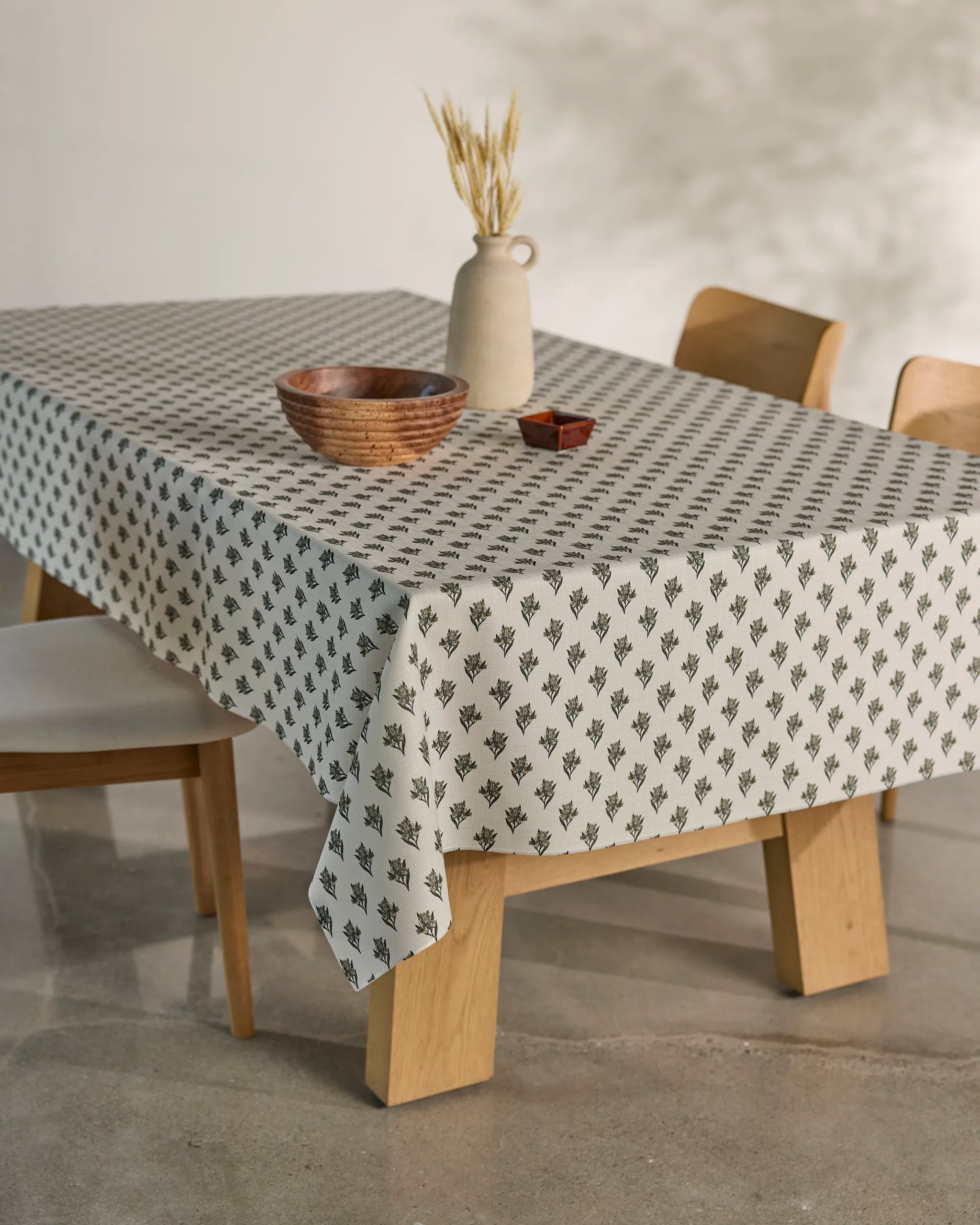 The Foraged tablecloth by Geometry, featuring a small black leaf pattern, covers a wooden dining table set with a wooden bowl, ceramic vase with dried wheat, and a small rectangular object. Three light wood chairs complete the scene.