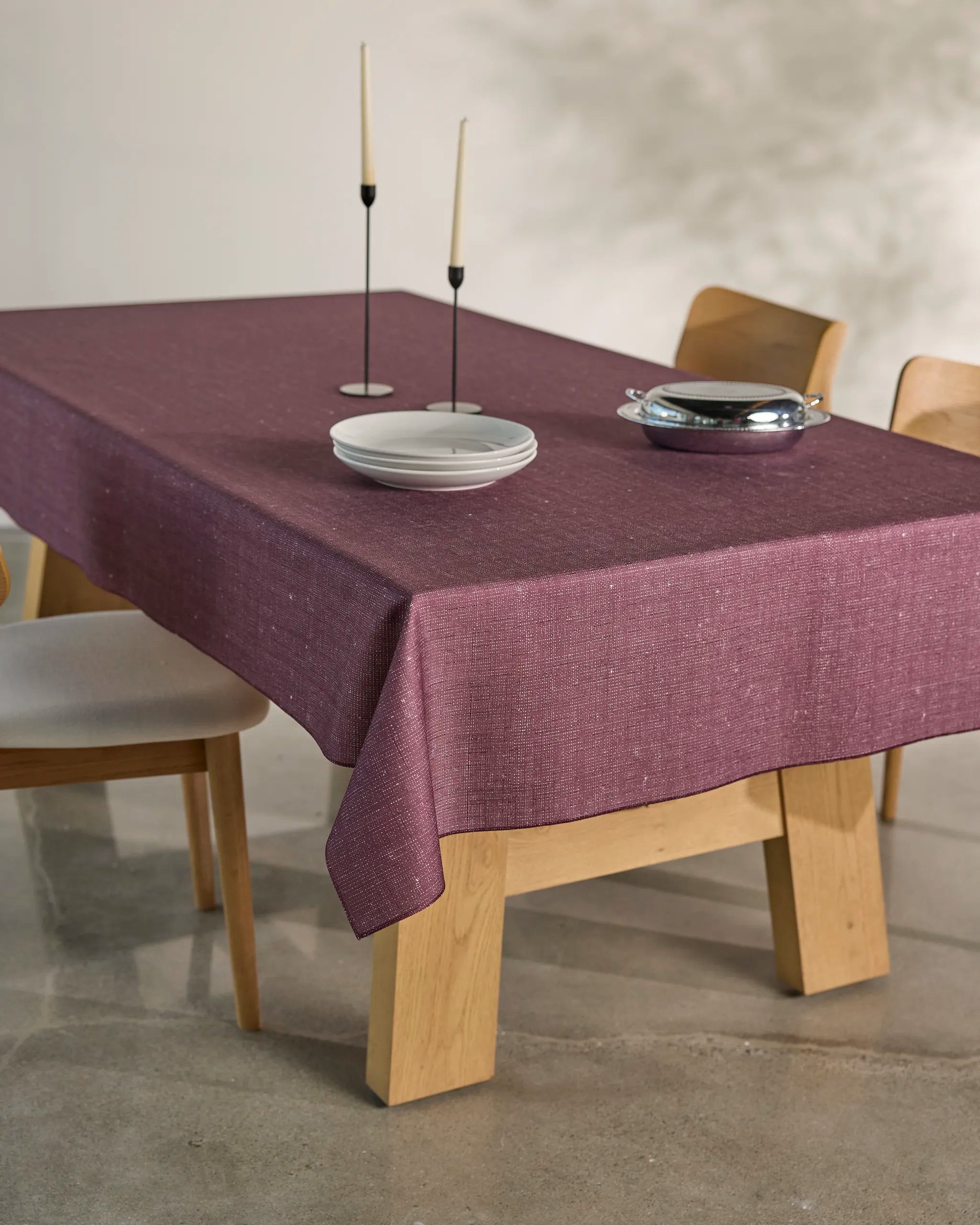 The Geometry Gather dining table features light wood and includes four matching chairs with white cushions. It's styled with a spill-resistant mauve tablecloth, stacked white plates, a silver dish, and two tall candles in black holders.