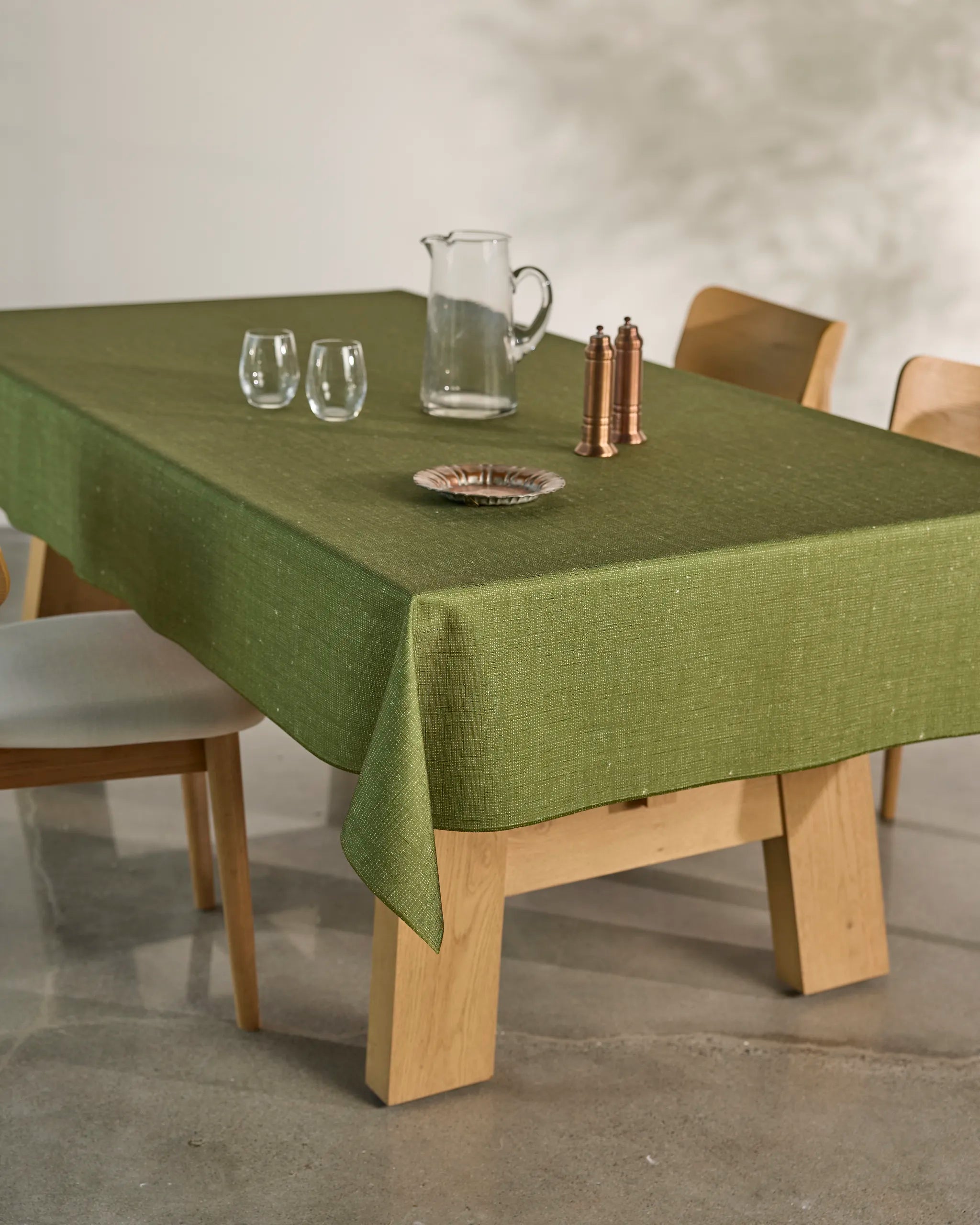 The Gather dining table by Geometry features a spill-resistant tablecloth, two clear glasses, a glass pitcher, copper salt and pepper shakers, and a small dish. Four light wood chairs complete the setting on a polished floor.