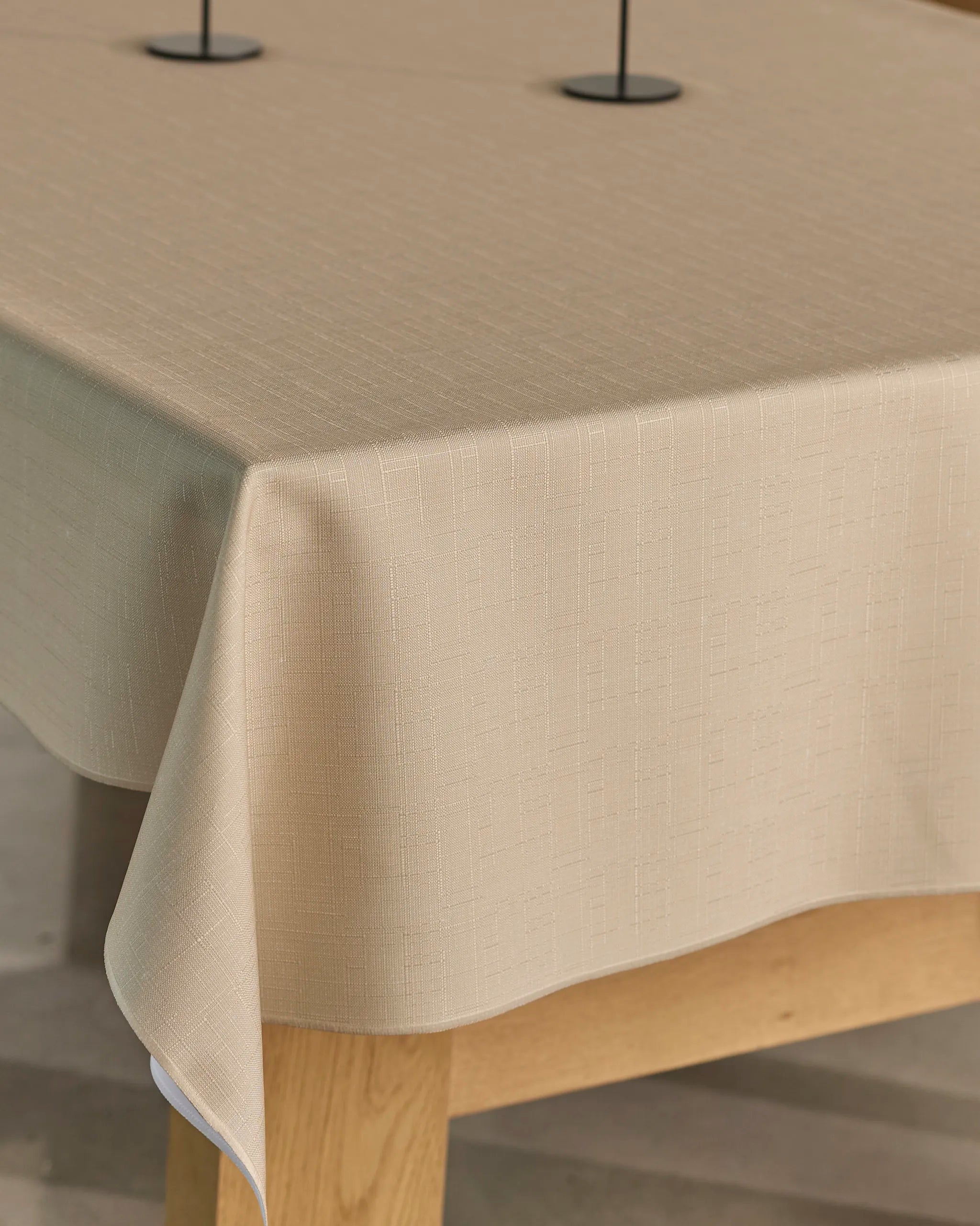 A close-up of the Geometry Gather tablecloth in beige, featuring a subtle textured pattern and spill-resistant fabric, draped neatly over a light wooden table with two thin-poled bases partly visible on top.