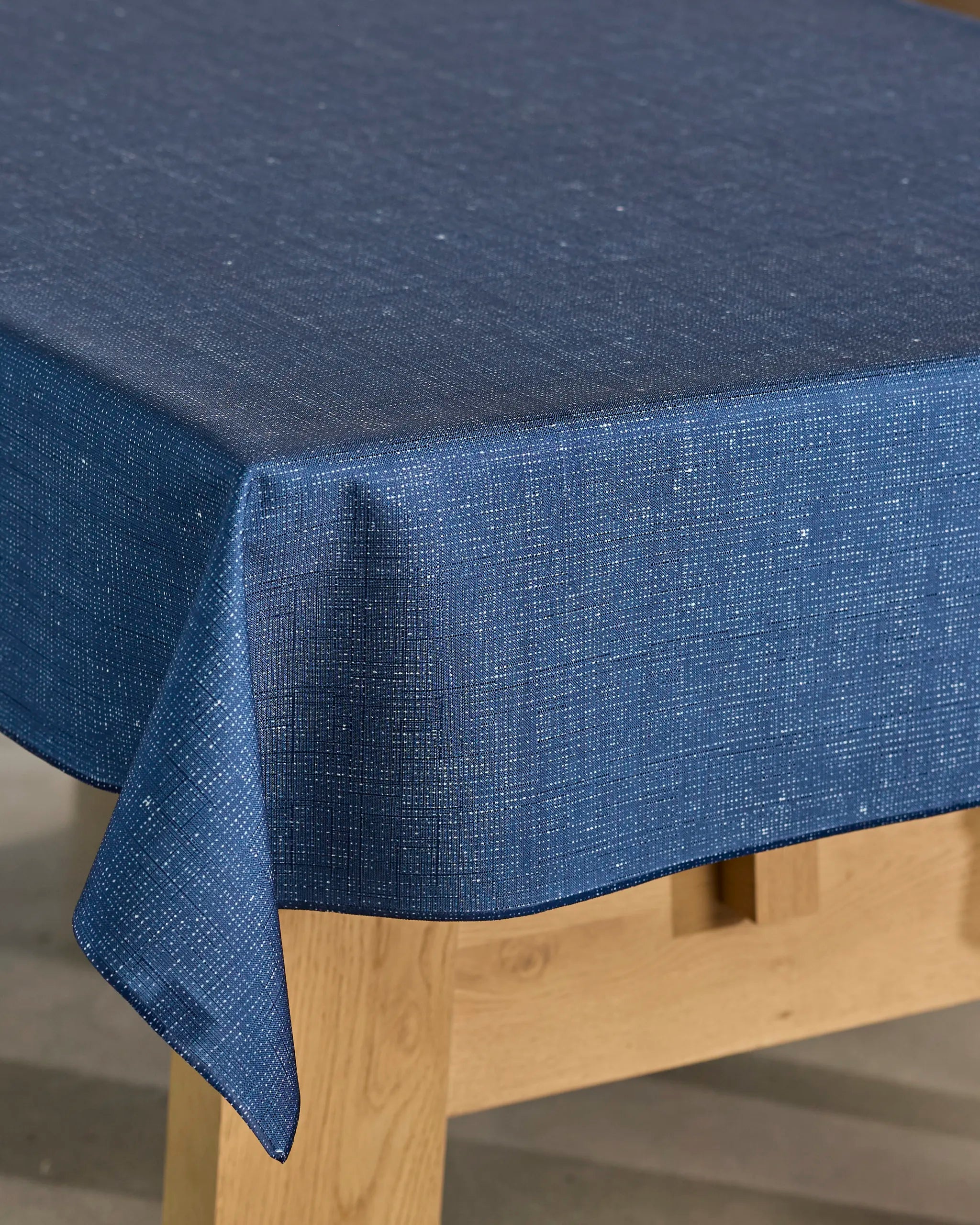 A close-up of Geometry's Gather tablecloth—a blue, spill-resistant cover with a subtle dotted pattern—draped over the edges of a rectangular wooden table.