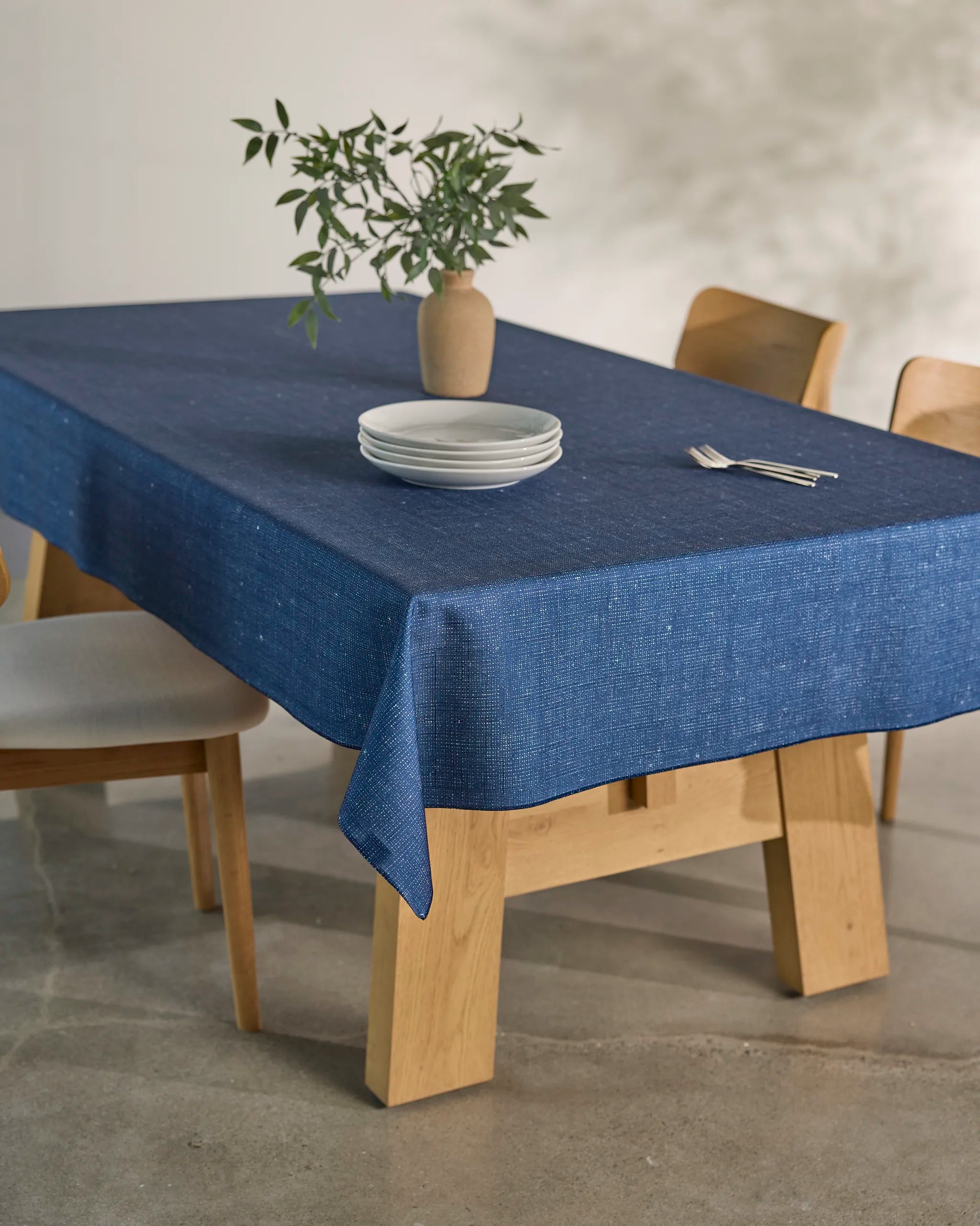 The Gather dining table by Geometry features a blue, spill-resistant tablecloth, a stack of white plates, silverware, and a beige vase with green branches. Four light wood chairs surround this elegant wooden table.