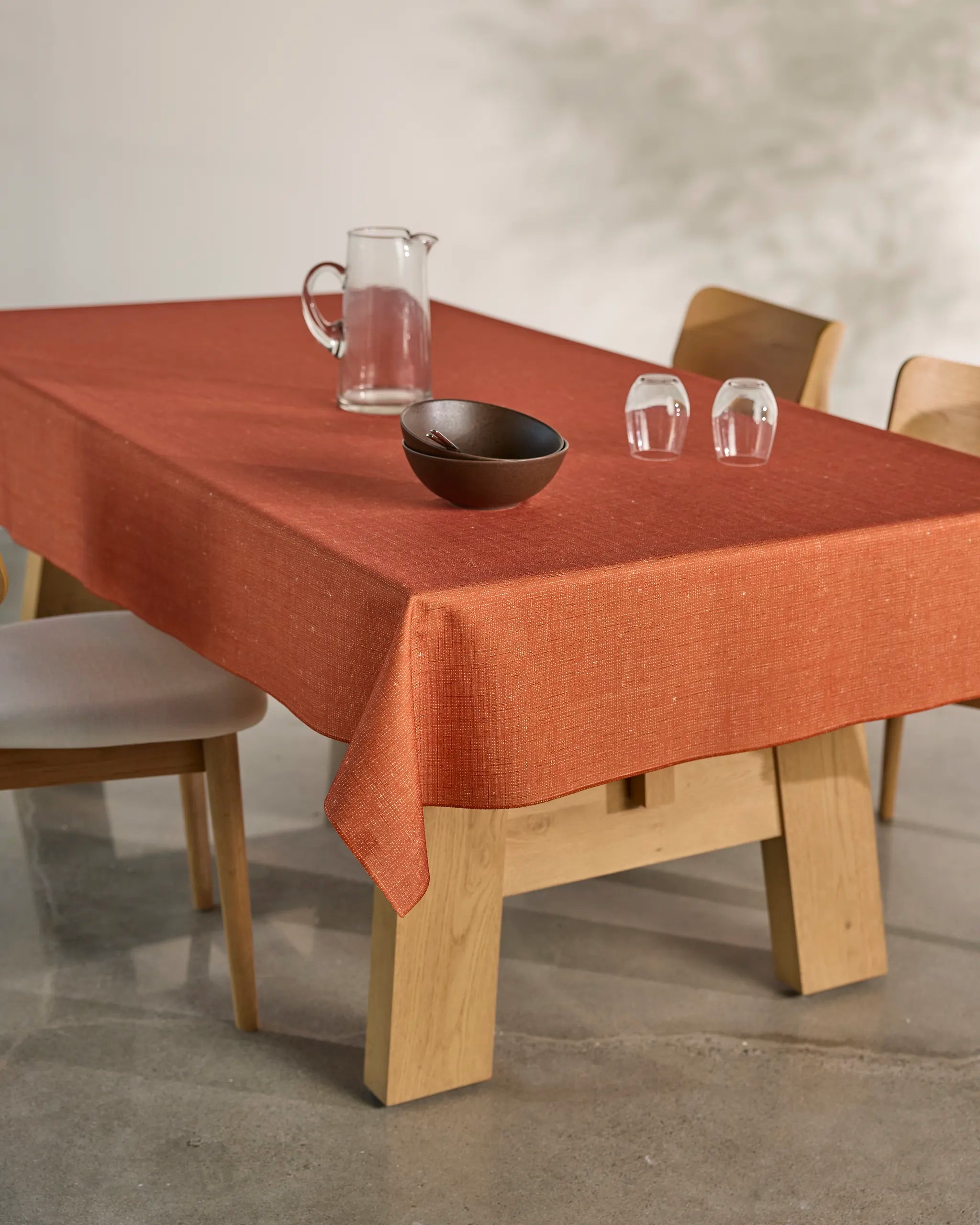 The Geometry Gather dining table features light wood surrounded by four matching chairs, topped with a spill-resistant burnt orange tablecloth, a clear glass pitcher, dark bowl, and two upside-down glasses on a concrete floor.