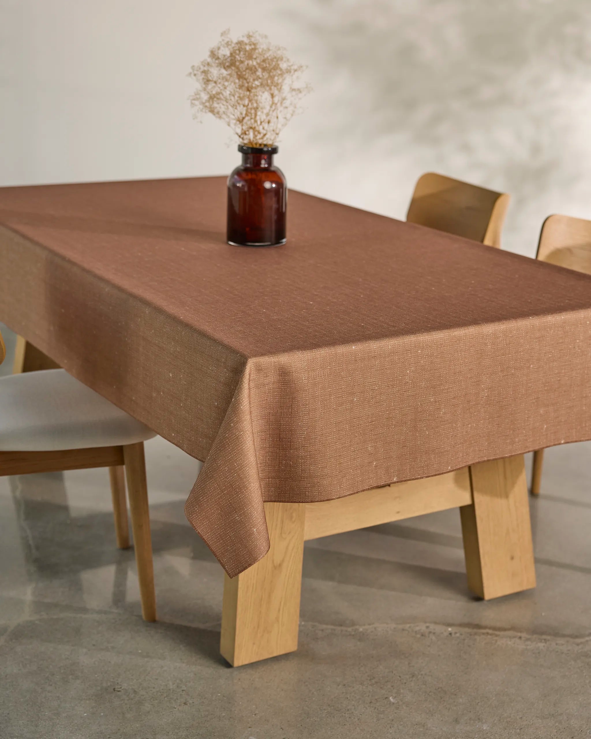 The Gather dining table by Geometry, topped with a spill-resistant tablecloth, is encircled by light wood chairs. At its center sits a brown glass vase with dried flowers, all set on a polished concrete floor.
