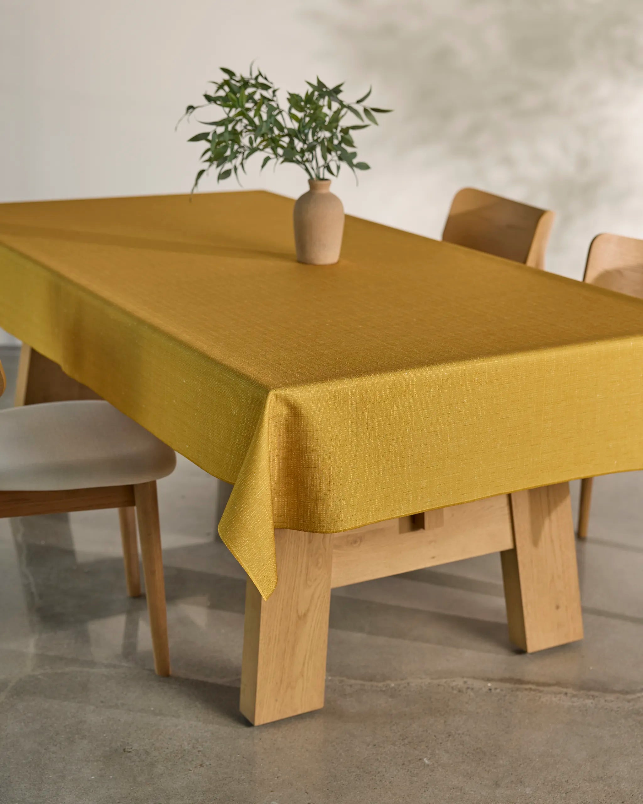 The Geometry Gather wooden dining table, draped in a yellow spill-resistant cloth and topped with a beige vase of green branches, is surrounded by wooden chairs on a polished floor.