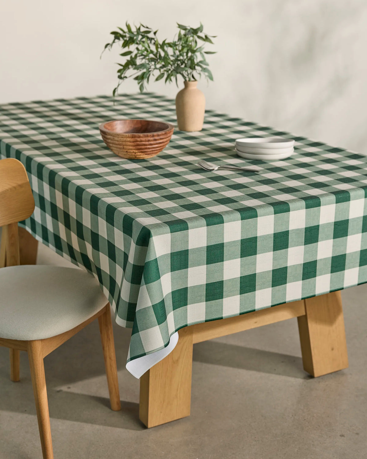 A wooden table is covered with the Geometry Holly Check spill-resistant green and white checkered tablecloth, set with a small vase of greenery, a wooden bowl, and stacked white plates. Beside the table sits a wooden chair with a cushioned seat.