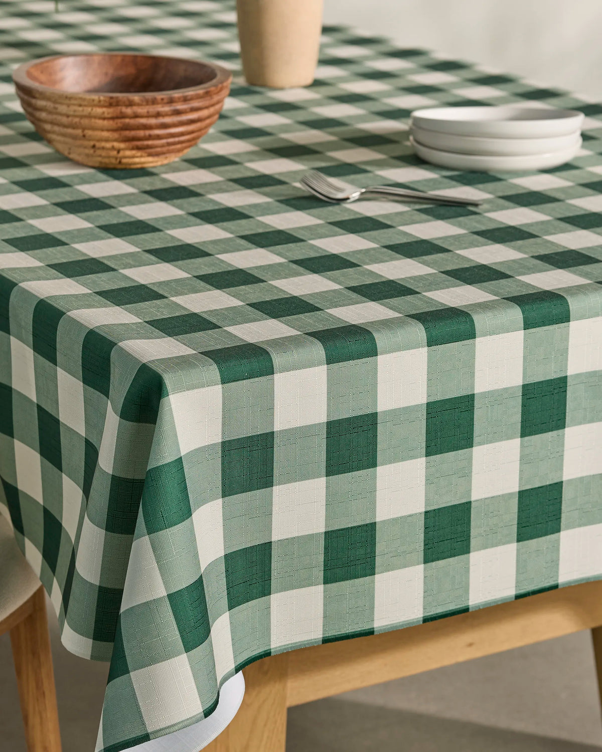 A wooden bowl, a small cup, and stacked white plates rest on a table covered with Geometry’s spill-resistant Holly Check green and white checkered tablecloth. The table has wooden legs and is set in a bright, minimalist room.