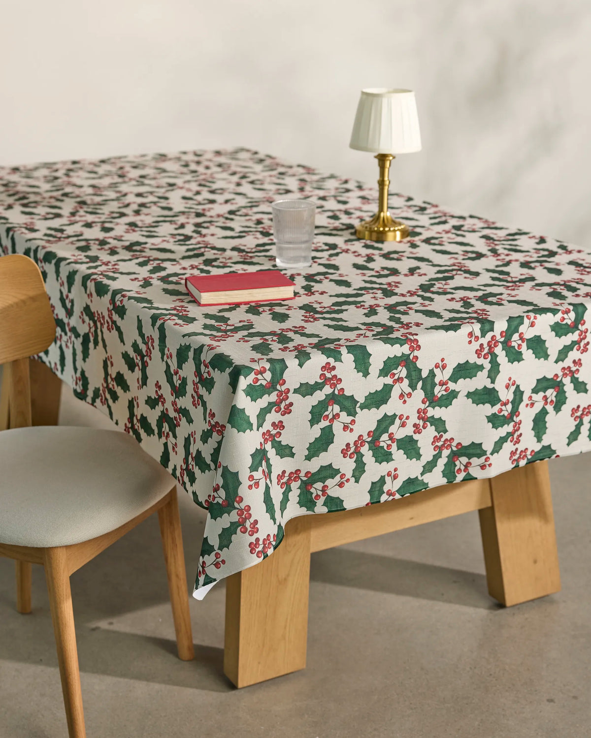 A wooden table is covered with Geometry’s “Holly Days” spill-resistant tablecloth, featuring a festive holly pattern. On the table are a glass of water, a red book, and a small lamp. Beside the table stands a wooden chair topped with a white cushion.