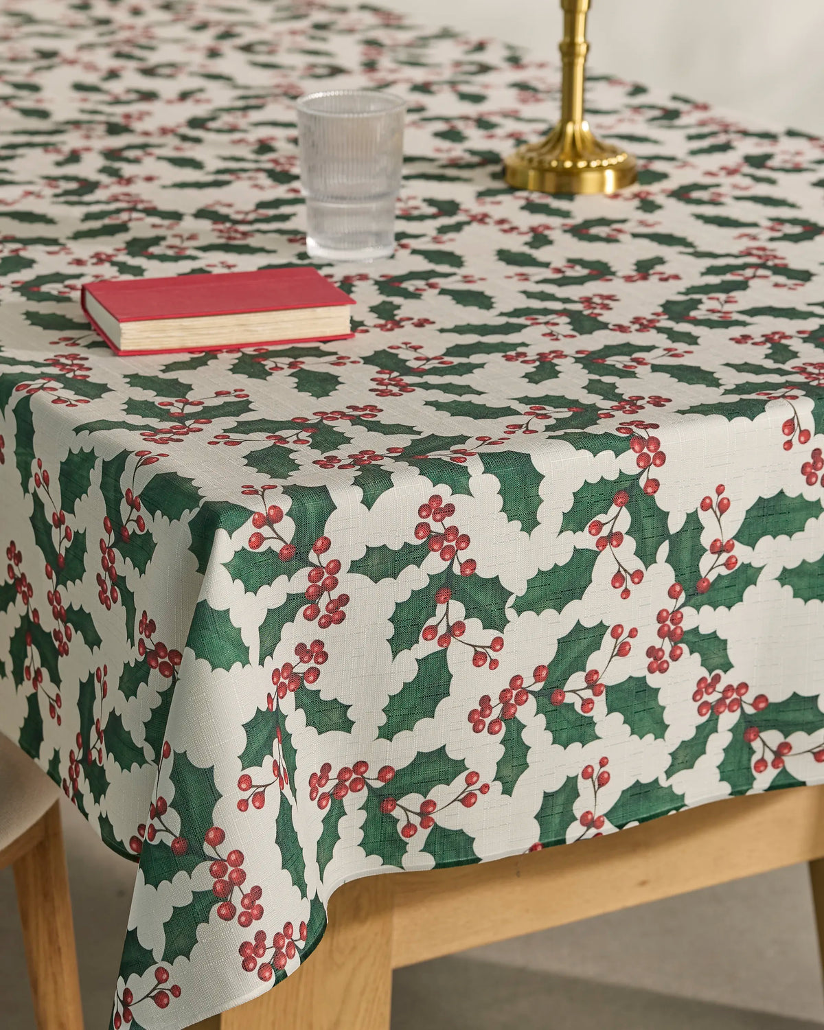 A table draped with Geometry’s “Holly Days” spill-resistant tablecloth holds a brass candlestick, a glass of water, and a closed red book.