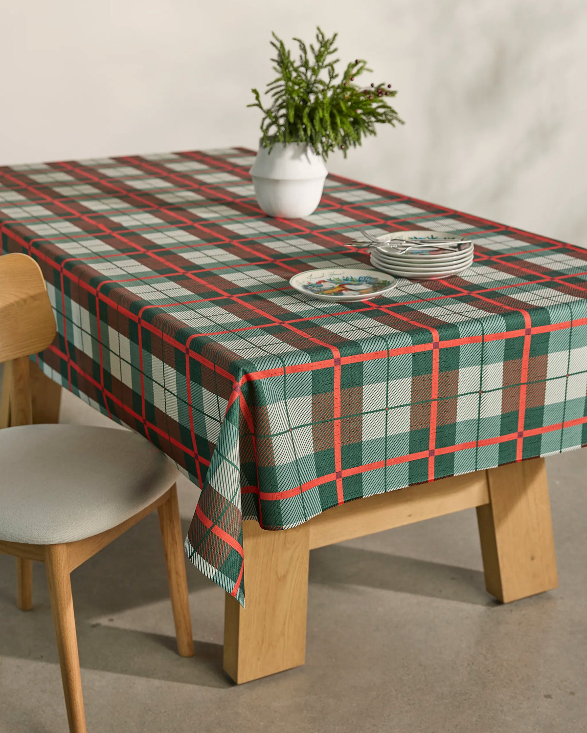 A wooden dining table featuring the Geometry Noel Tartan spill-resistant tablecloth in red, green, and white plaid is topped with a white vase filled with greenery and a stack of plates. Beside the table sits a cushioned wooden chair on the concrete floor.