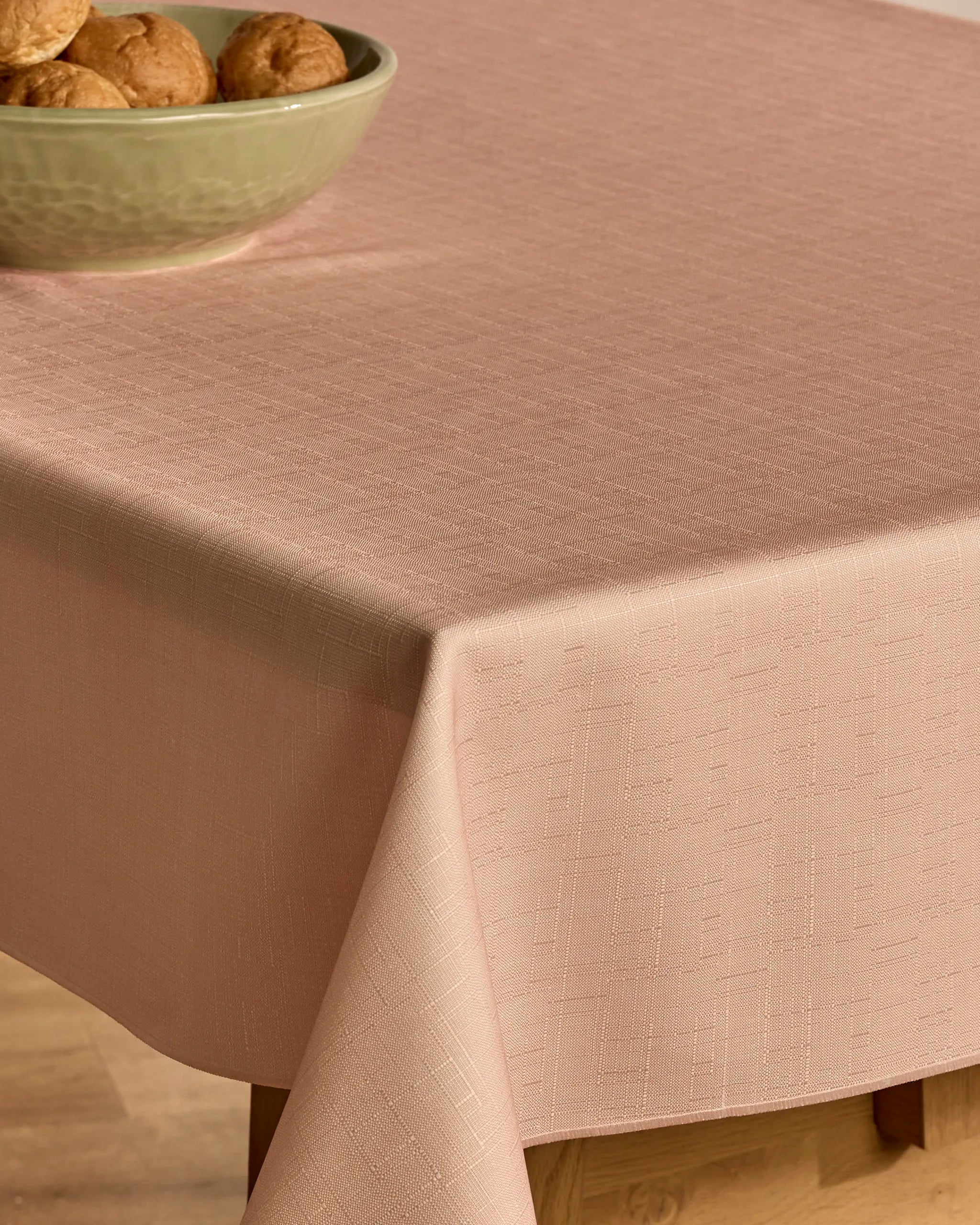 The Geometry Solid tablecloth in beige, designed for spill resistance, covers a light wood rectangular table. A green bowl of bread rolls is placed on the upper left corner of the table.