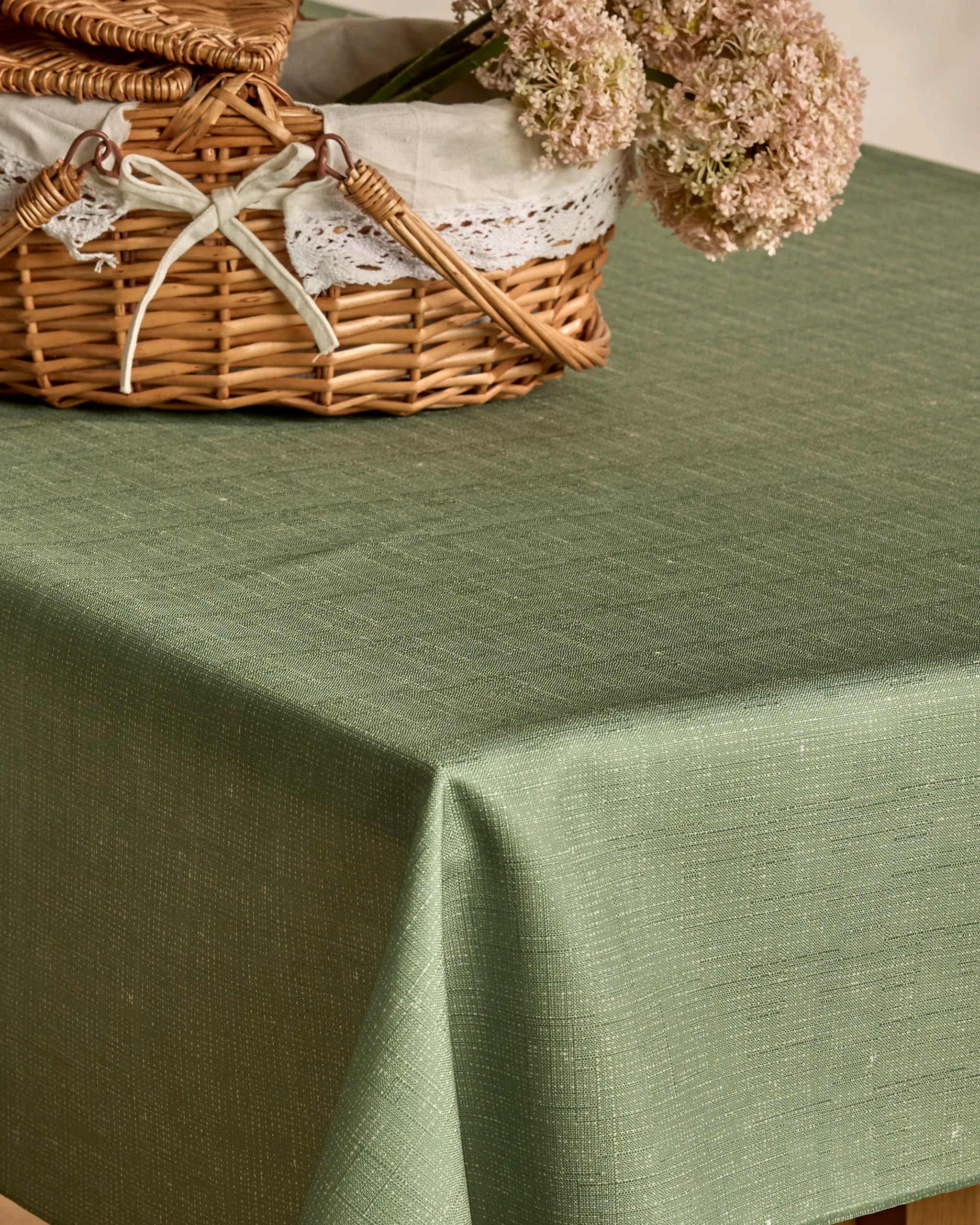 A Geometry Solid green lightweight tablecloth covers the table. On top is a wicker basket with lace trim and a white ribbon, placed beside pale pink flowers.
