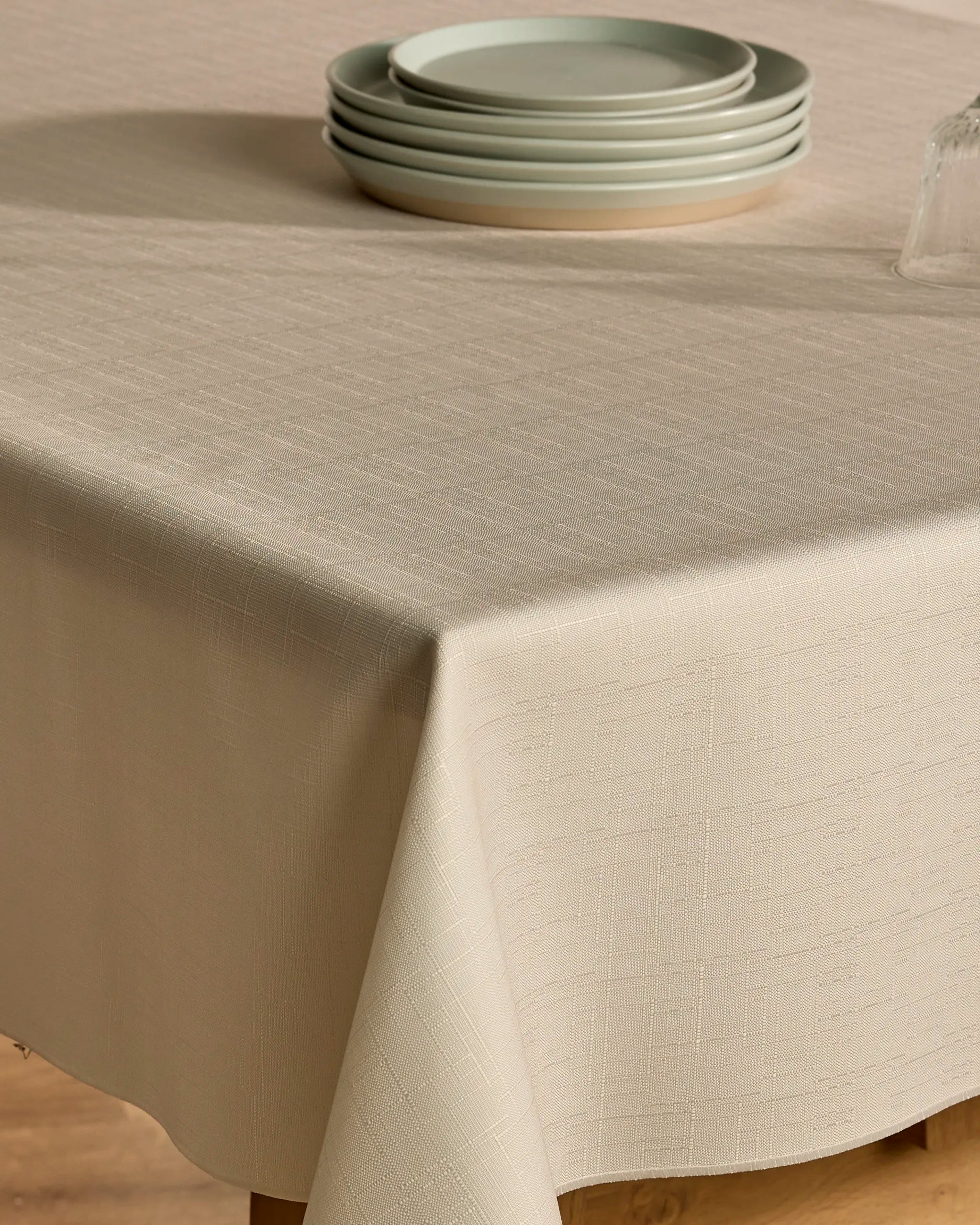 A close-up of the lightweight, beige "Solid" tablecloth by Geometry covers a table, topped with four stacked light gray plates and part of an upside-down glass. The tablecloth features a subtle textured pattern.