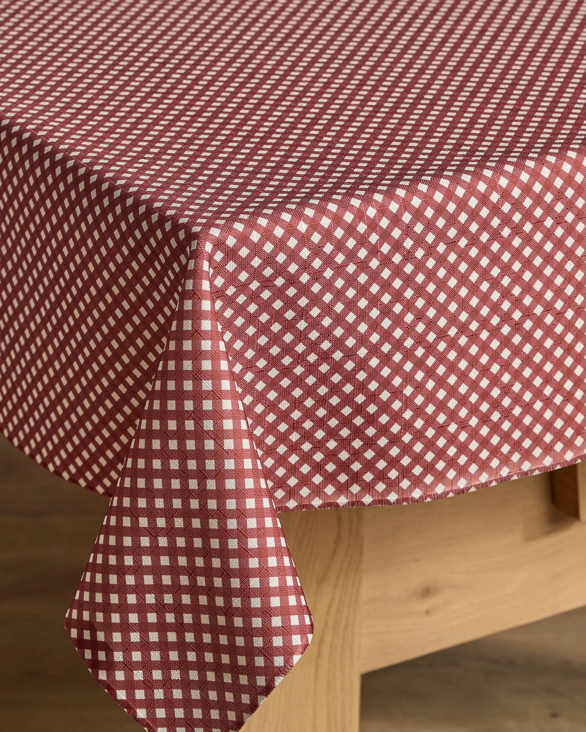 A round wooden table is adorned with the Bramble Red tablecloth by Geometry, featuring a stain-resistant, red and white diamond pattern that drapes elegantly over the edges, complementing the warm-toned floor beneath.