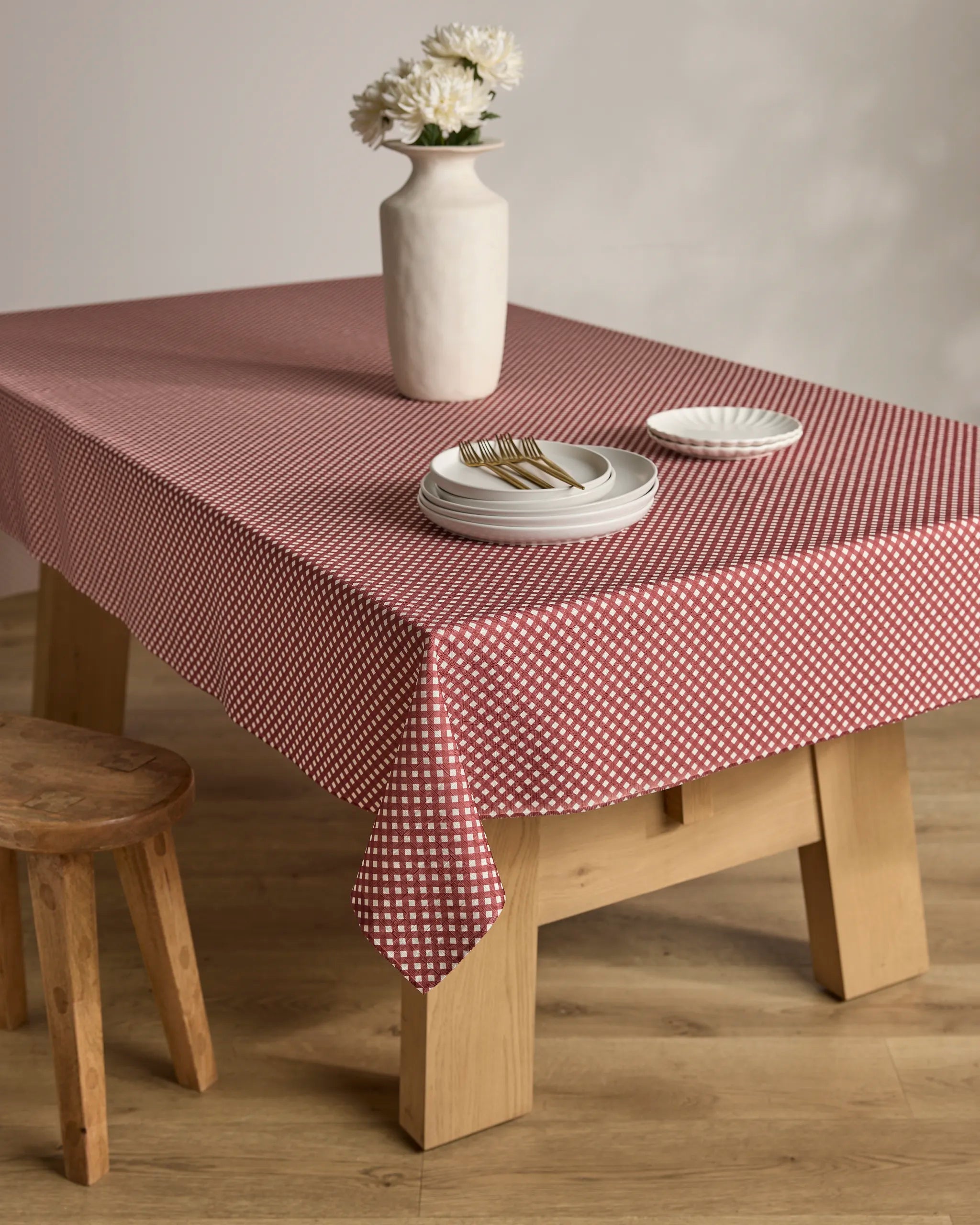 A wooden table with the Bramble Red stain-resistant, red and white checkered tablecloth by Geometry is set with stacked white plates, forks, a small dish, and a white vase with flowers. A wooden stool sits beside the table.