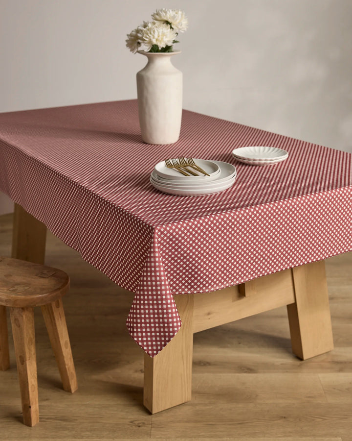A wooden table with the Bramble Red stain-resistant, red and white checkered tablecloth by Geometry is set with stacked white plates, forks, a small dish, and a white vase with flowers. A wooden stool sits beside the table.
