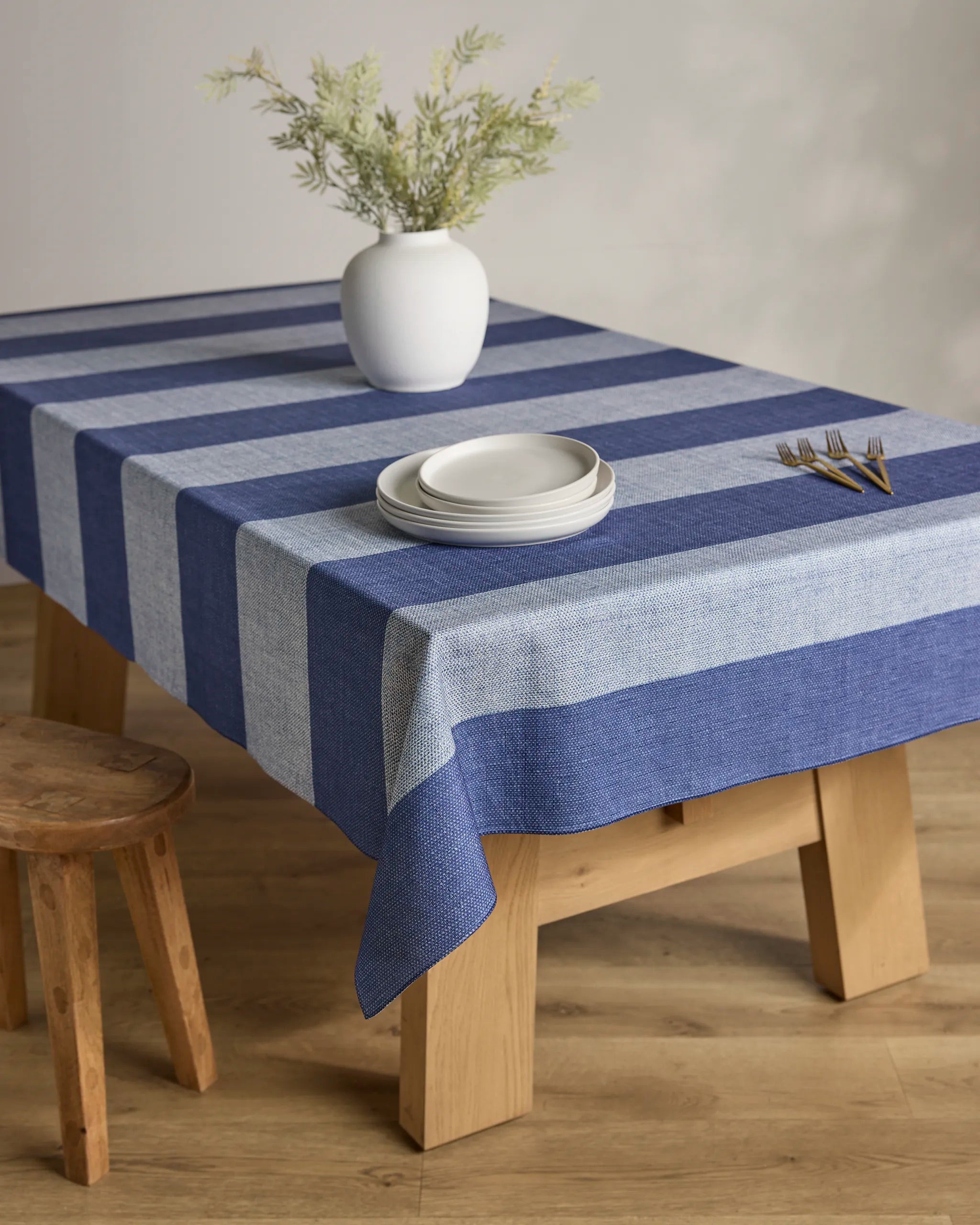 A wooden table covered with the Geometry Everband blue and gray striped stain-resistant tablecloth is set with white plates, four forks, a white vase with greenery, and a wooden stool, all on a light wood floor.