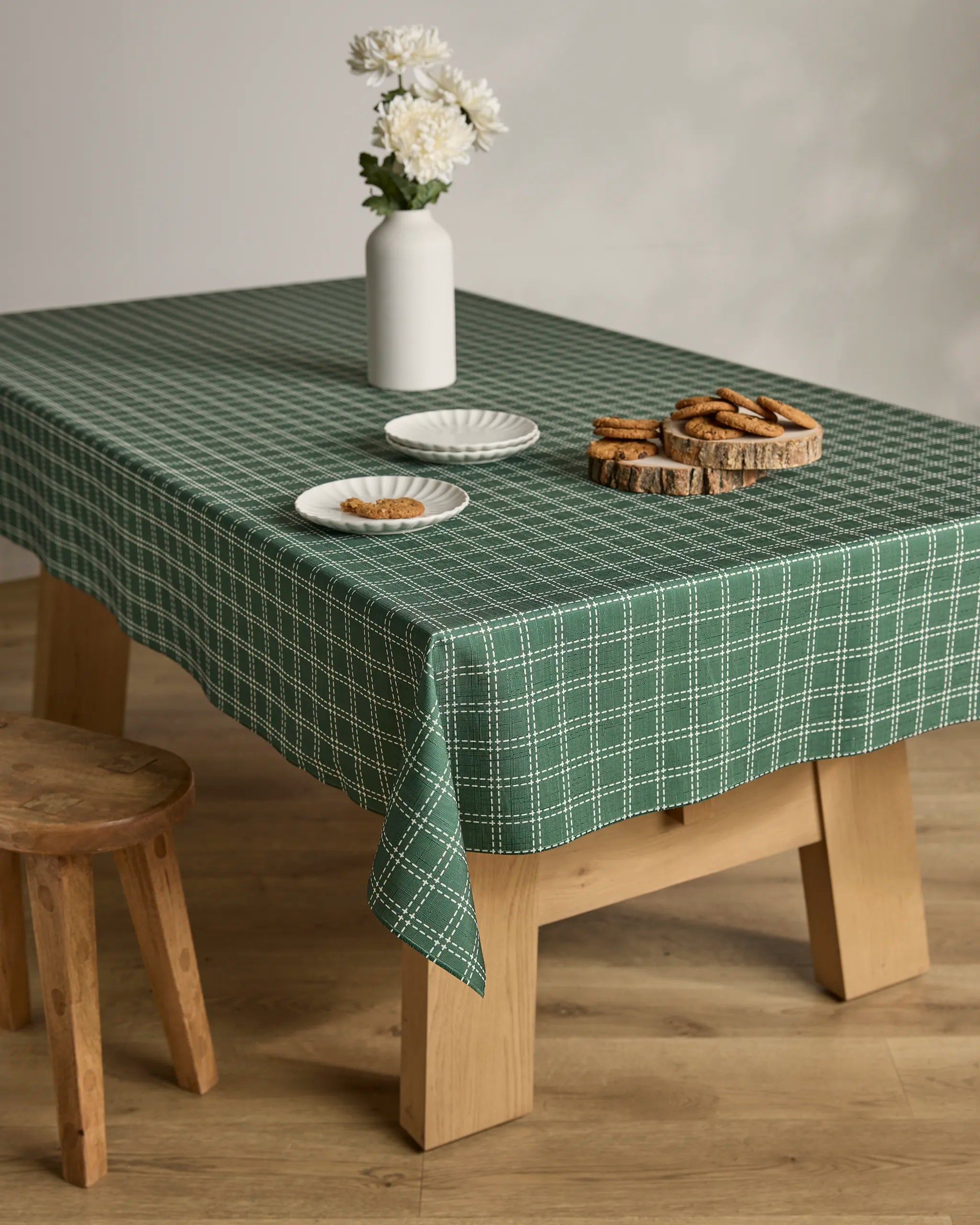 A Geometry Firwood table covered with a stain-resistant green and white checkered cloth displays a vase of white flowers, two plates of cookies, and a wooden platter with more cookies. A small wooden stool sits nearby.