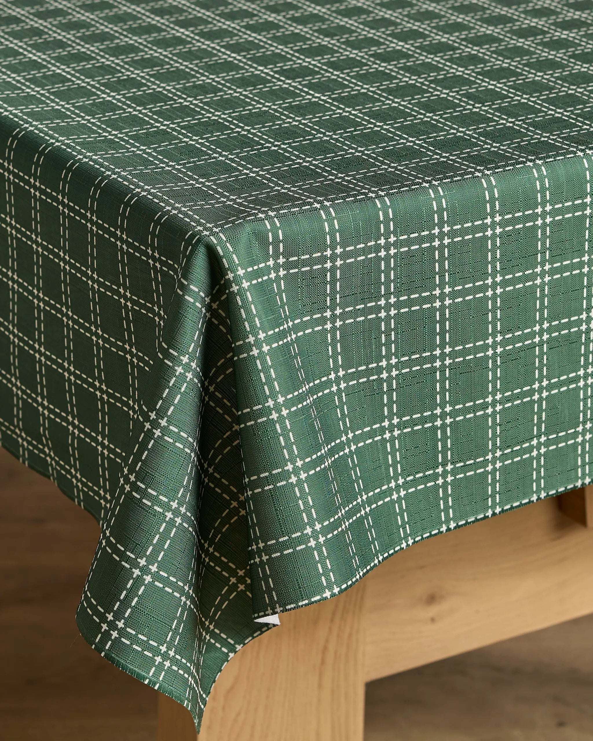 The Geometry Firwood tablecloth, featuring a lightweight green fabric with a white plaid design, drapes neatly over the corner of a light wooden table.