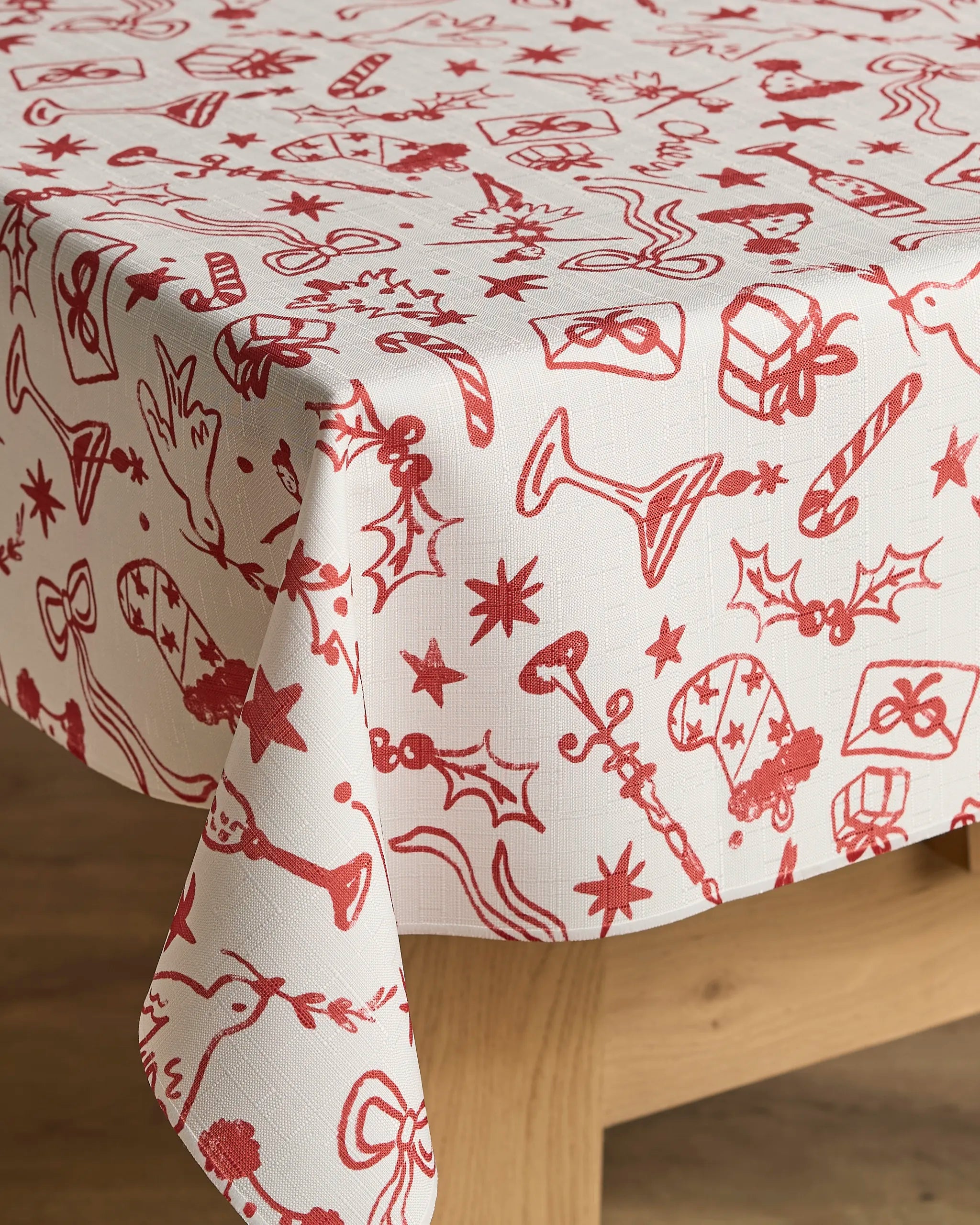 The Geometry Holiday Doodles tablecloth, featuring red festive sketches, drapes elegantly over a wooden table, adding holiday charm with its stain-resistant and easy-to-clean design.