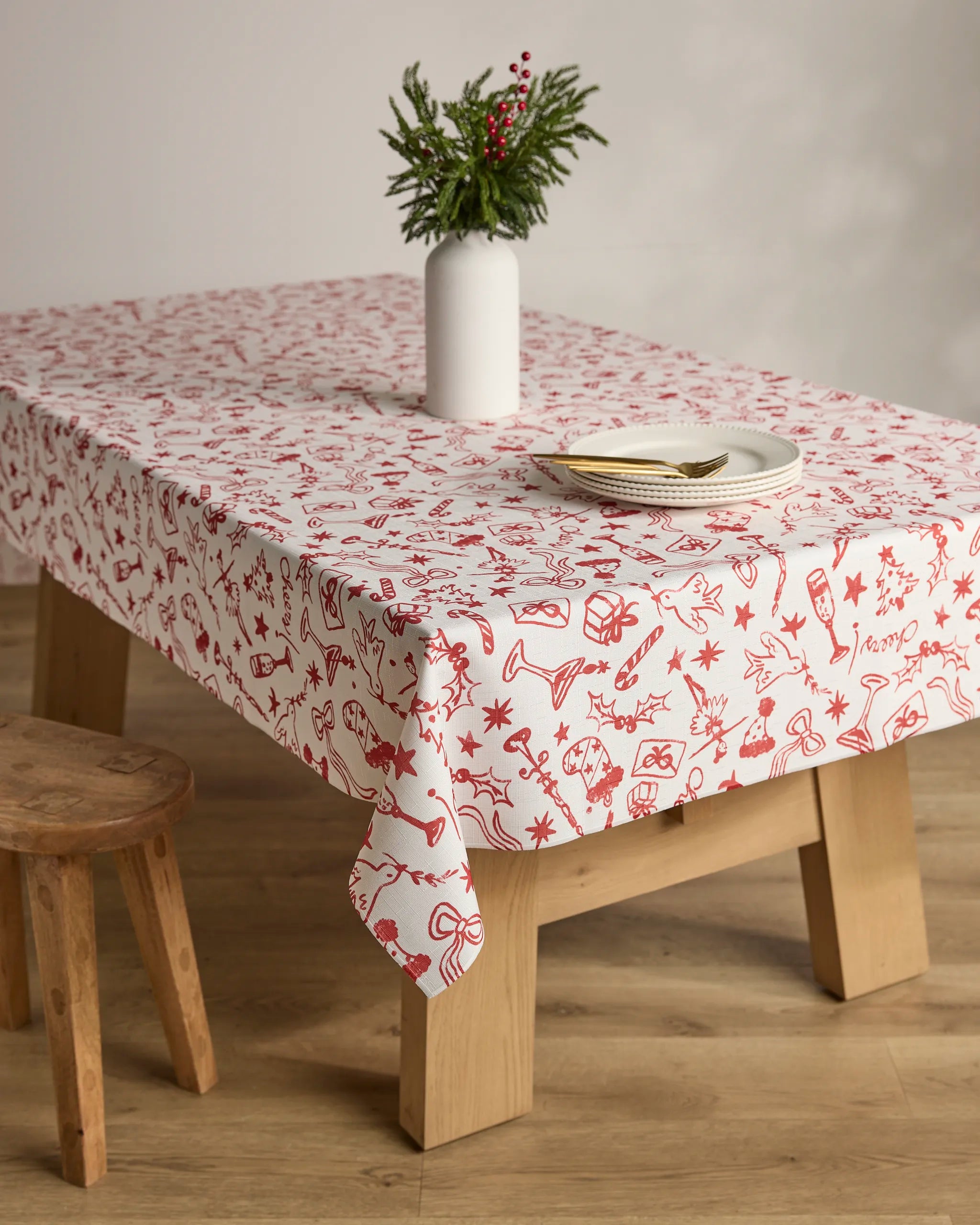 A Geometry "Holiday Doodles" stain-resistant red-and-white tablecloth covers a wooden table set with two plates, cutlery, and a white vase with green foliage and red berries; a small wooden stool sits beside it on a wooden floor.