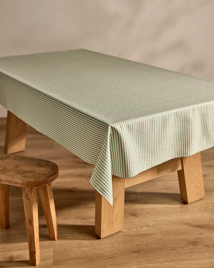 A rectangular wooden table is adorned with the Kailee Wright Mint Green Stripe eco-friendly tablecloth, paired with a wooden stool on a wooden floor and set against a neutral wall.